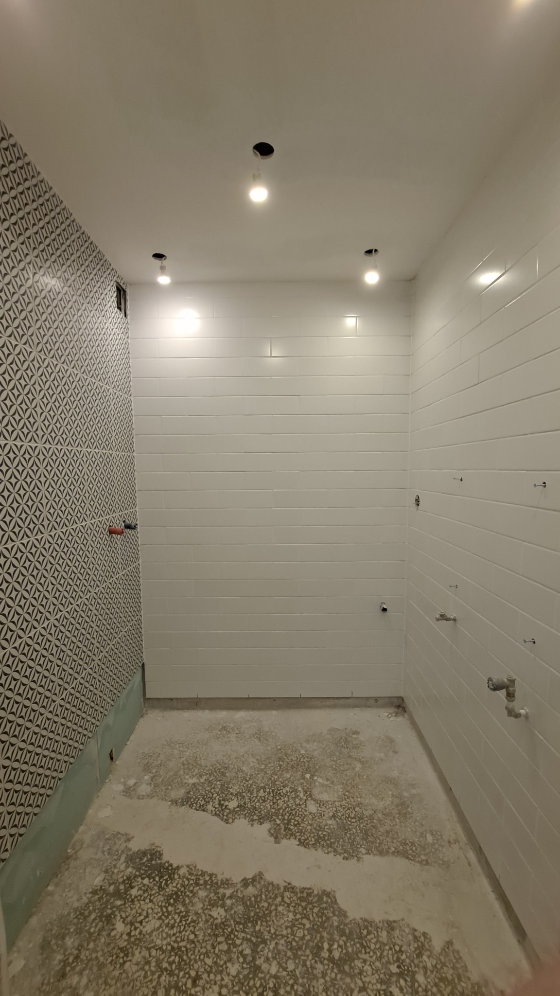 Wnętrze łazienki w trakcie remontu: ściany częściowo wyłożone białymi, podłużnymi płytkami, jedna ściana z wzorzystą tapetą, surowa podłoga, widoczne punkty oświetleniowe w suficie i instalacje...