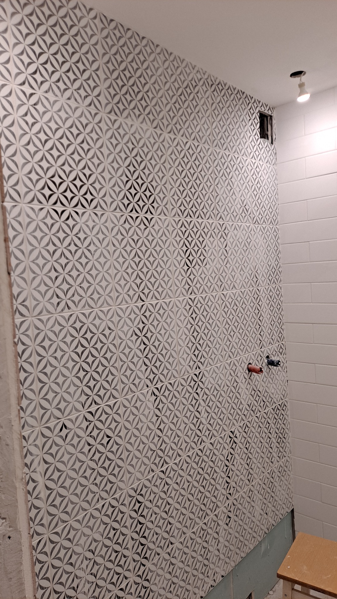 Ściana łazienki wyłożona wzorzystymi, jasnymi płytkami ceramicznymi z ciemnoszarym motywem kwiatowym, widoczne wystające rury instalacji wodnej i fragment ściany z białymi, prostokątnymi płytkami.