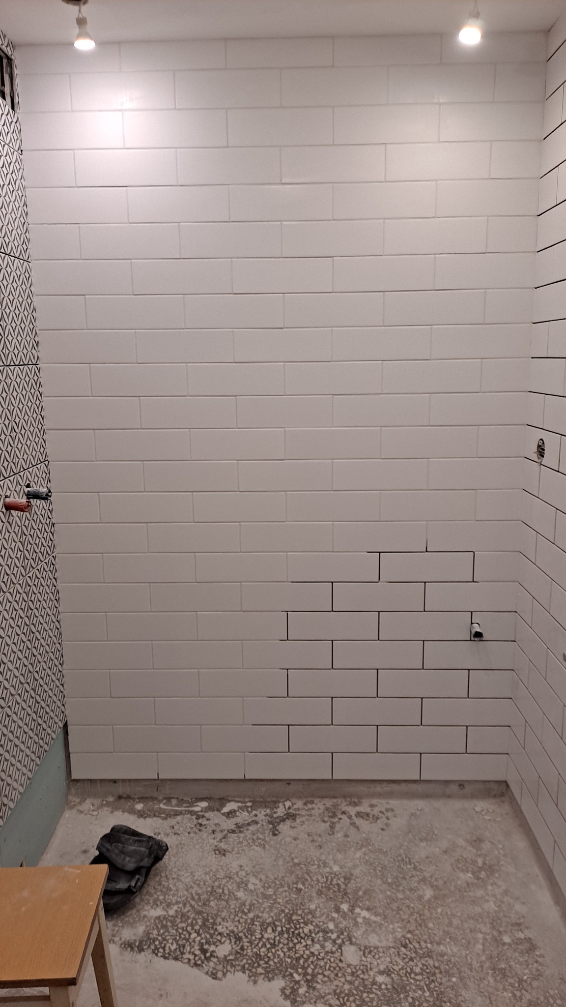 Ściana łazienki w trakcie układania białych, podłużnych płytek; widoczne niedokończone fragmenty i wystające rury; na podłodze resztki zaprawy i szary worek; w tle fragment ściany z wzorzystymi...