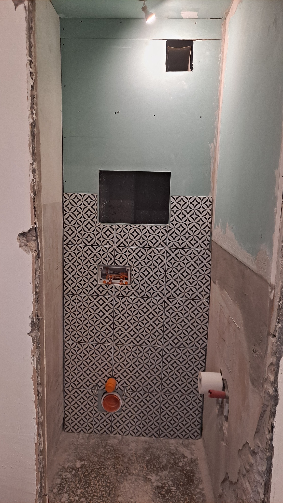 Ściana w trakcie remontu łazienki z częściowo ułożonymi czarno-białymi płytkami o wzorze geometrycznym, widoczne instalacje hydrauliczne i niedokończone ściany z płyt gipsowo-kartonowych.