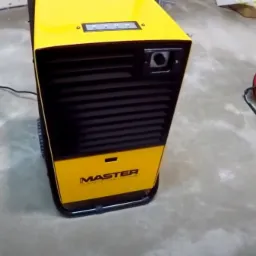 Żółto-czarny osuszacz powietrza marki Master z widocznym panelem sterowania i wtyczką, ustawiony na szarej, betonowej podłodze. W tle widoczny czerwony wentylator.