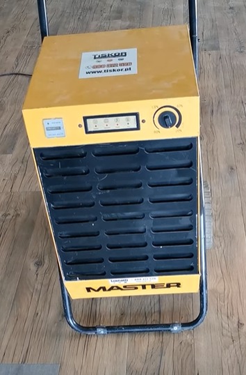 Żółty, przenośny osuszacz powietrza marki Master, z widocznymi panelami sterowania i napisem Tiskor na obudowie, stojący na drewnianej podłodze.
