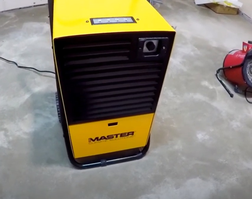 Żółto-czarny osuszacz powietrza marki Master z widocznym panelem sterowania i wtyczką, ustawiony na szarej, betonowej podłodze. W tle widoczny czerwony wentylator.