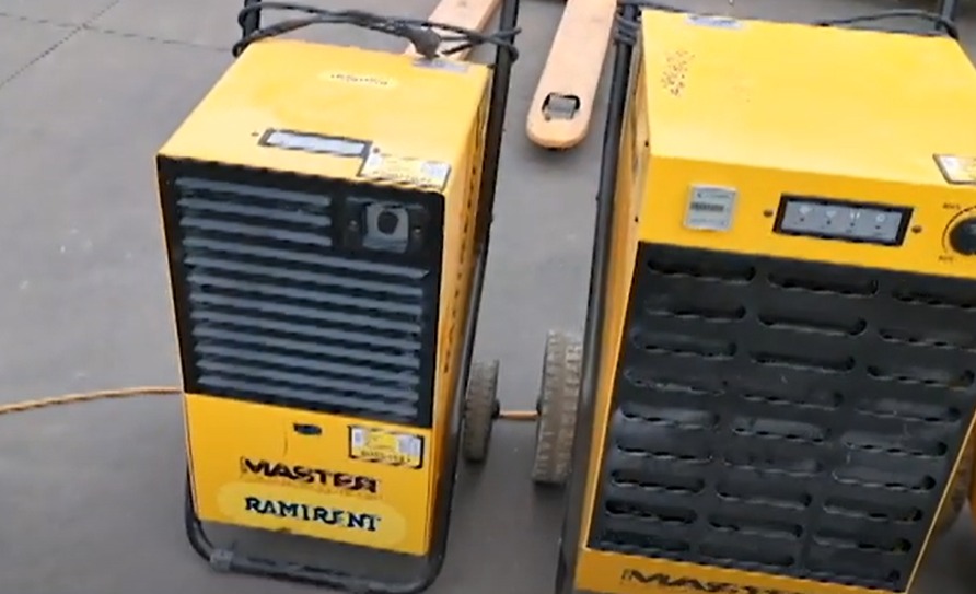 Dwa żółte, mobilne osuszacze powietrza marki Master Ramirent na szarym tle podłogi, widoczne przewody i kółka.