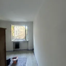 Wnętrze pokoju po malowaniu na biało, z widocznym oknem i grzejnikiem. Na podłodze wiadro z farbą i folia ochronna. Świeżo odmalowane ściany z niebieską taśmą malarską.