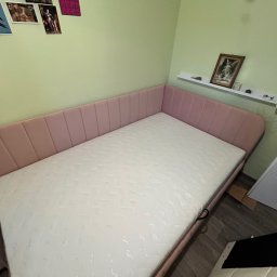 Patryk Milczanowski - Różowe łóżko z materacem w rogu pokoju o pistacjowych ścianach. Nad łóżkiem półka z dekoracjami i obrazem. Na ścianie wiszą grafiki.