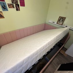 Patryk Milczanowski - Różowe łóżko tapicerowane z białym materacem w pokoju z zielonymi ścianami. Na ścianie wiszą plakaty i obrazek w ramce na półce.