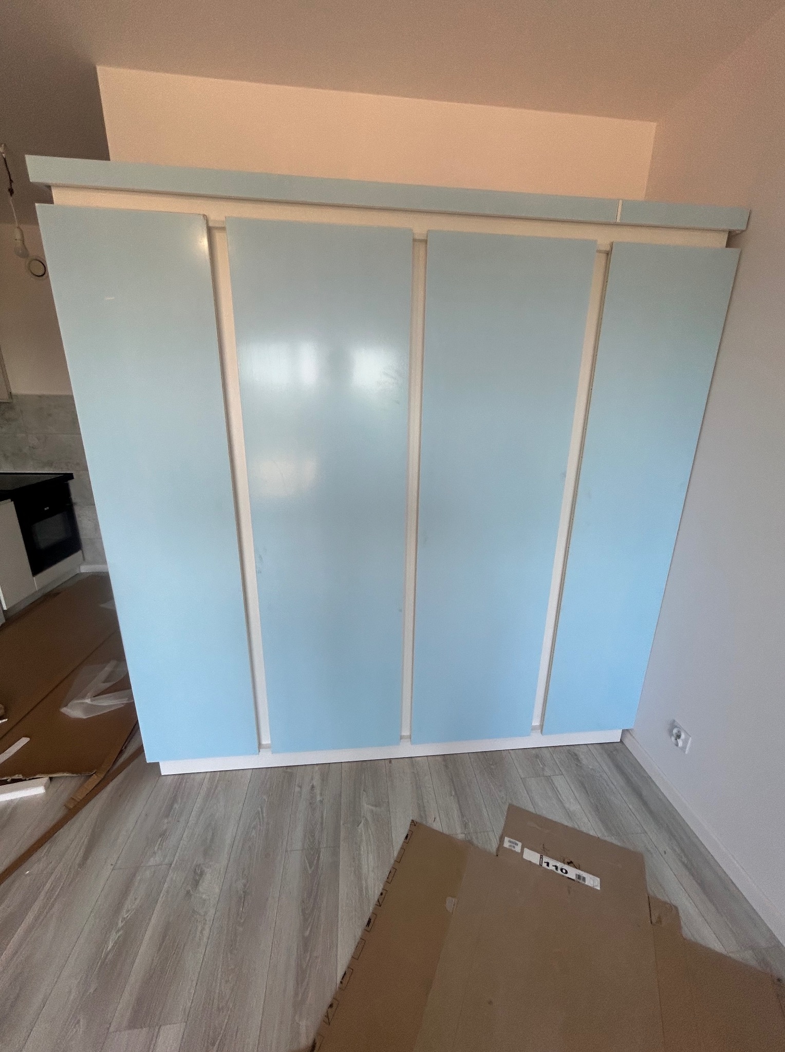 Świeżo zamontowana szafa z czterema przesuwnymi, błękitnymi drzwiami w nowoczesnym wnętrzu. Na podłodze widoczne kartony po meblach i panele podłogowe.