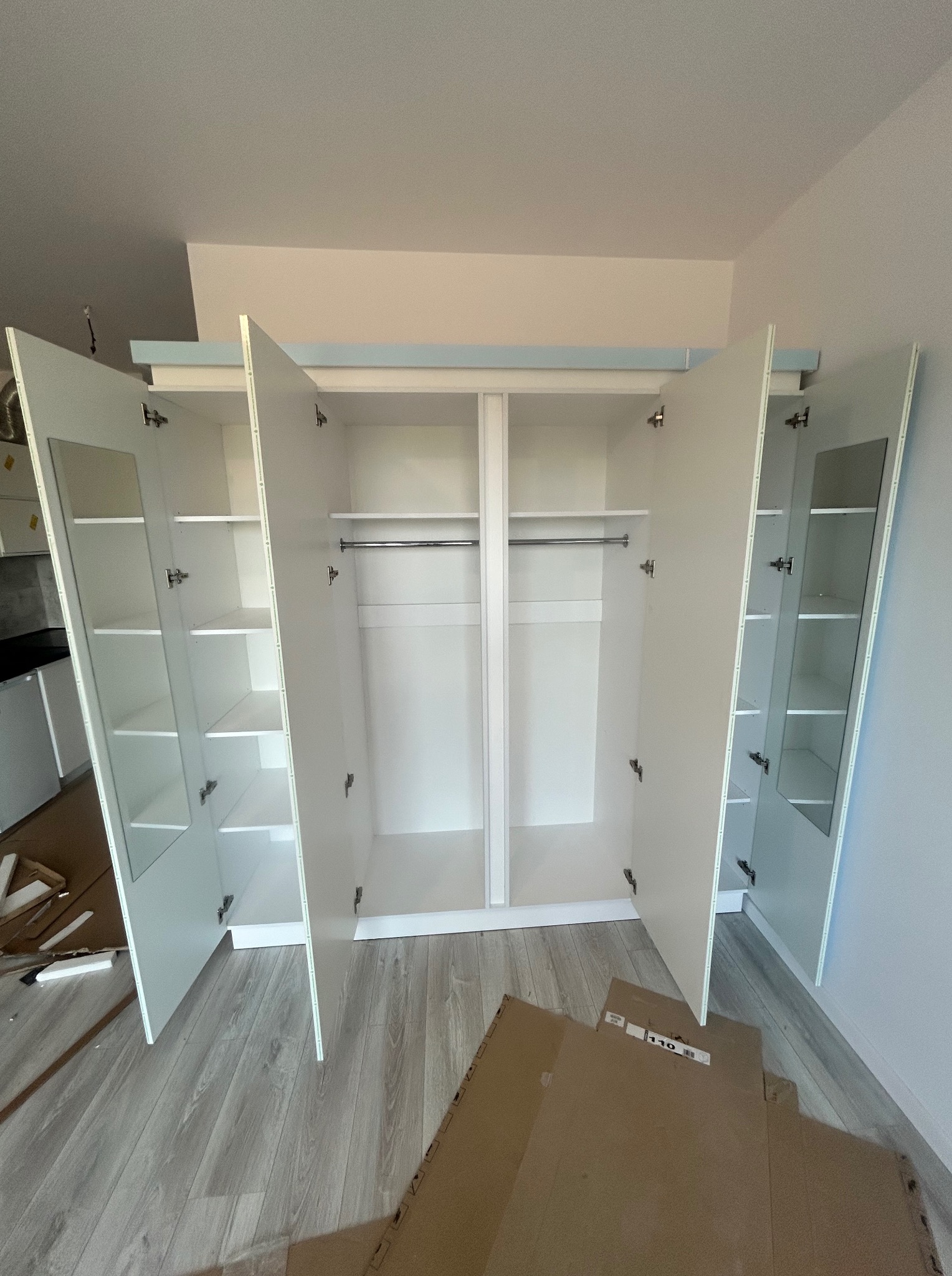Biała, trzydrzwiowa szafa z lustrami po bokach, z otwartymi drzwiami, ukazująca puste półki i drążki na ubrania, z kartonami na podłodze. Nowoczesny design.