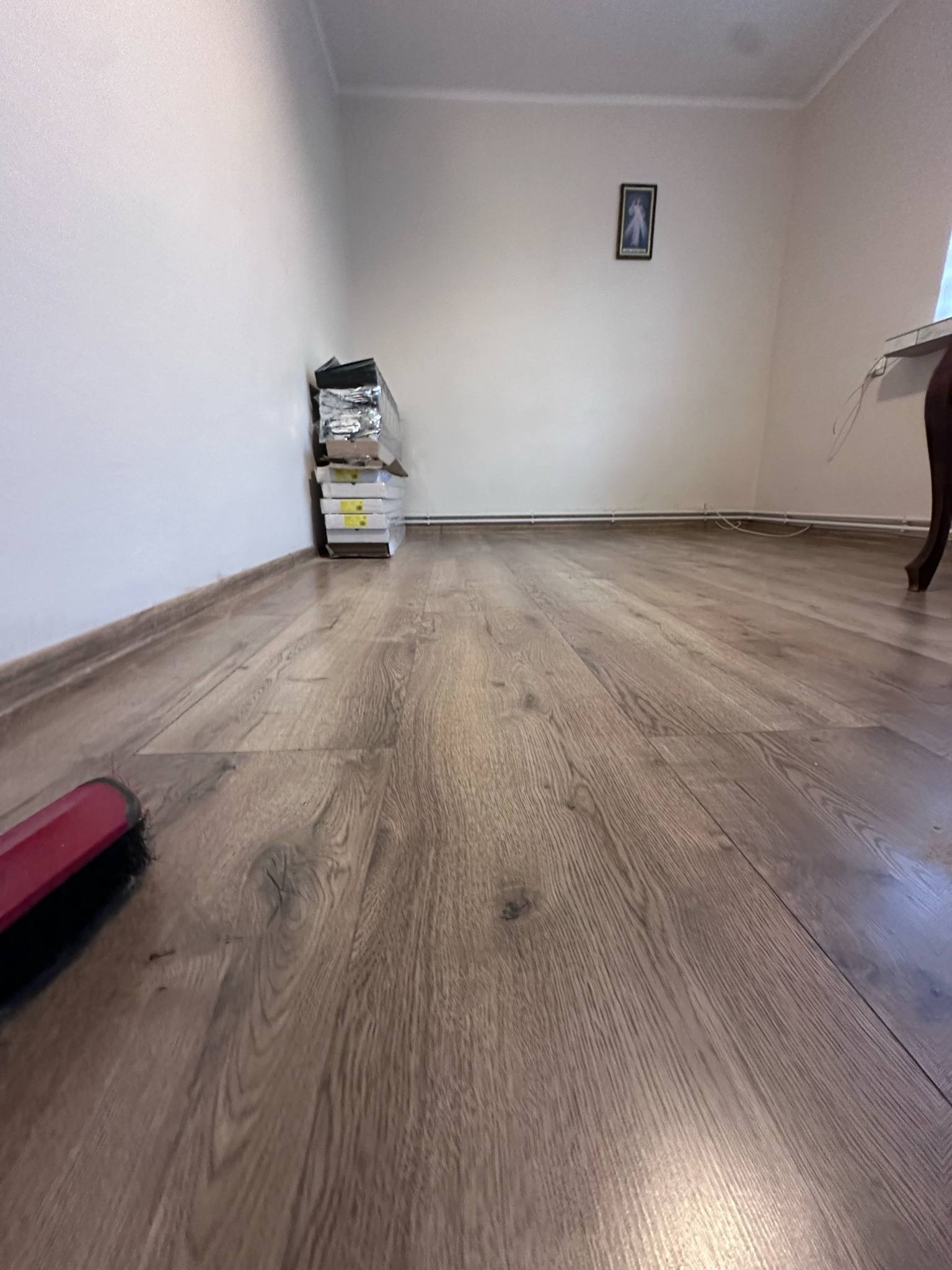 Ułożone panele podłogowe w pokoju, widoczne paczki paneli przy ścianie i fragment szczotki do sprzątania, ciepłe oświetlenie.