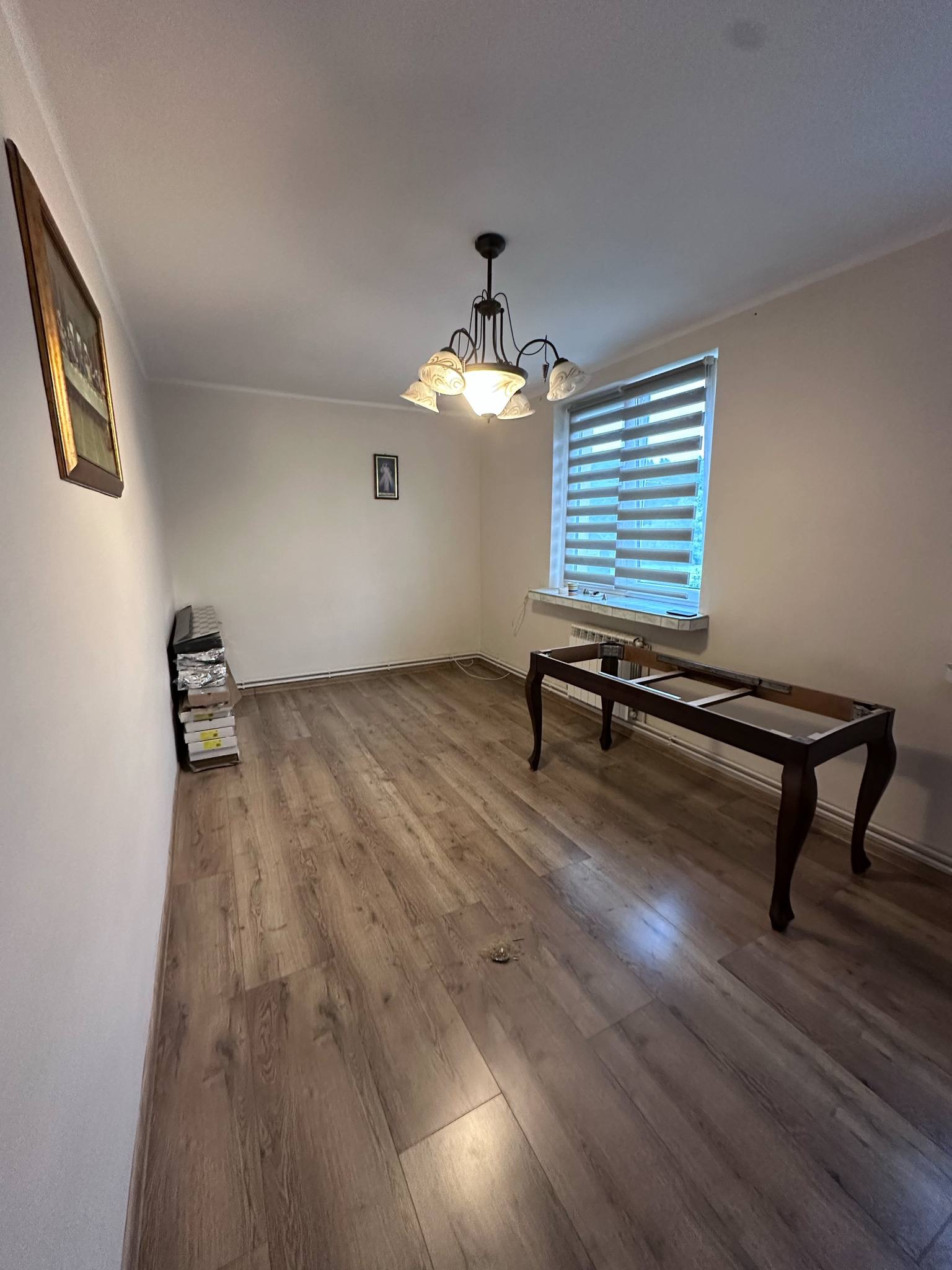 Pokój z drewnianą podłogą, oświetlony żyrandolem. Widoczne okno z żaluzjami, obraz na ścianie i częściowo rozłożony stół. Przy ścianie ułożone materiały budowlane.