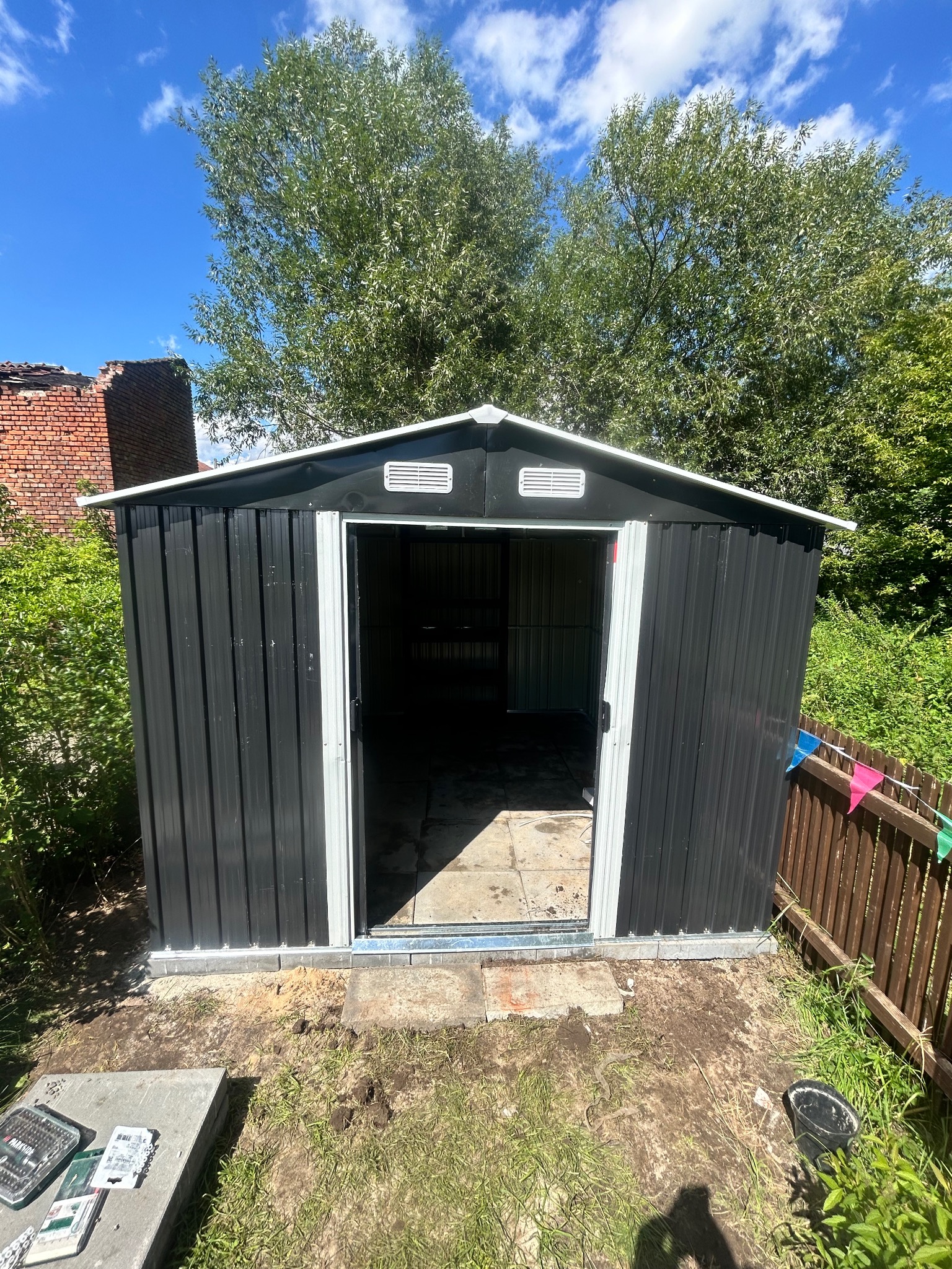 Zmontowany, metalowy domek narzędziowy w ogrodzie, z otwartymi drzwiami. Widoczne narzędzia obok, ceglany budynek w tle i drzewa. Słoneczny dzień.