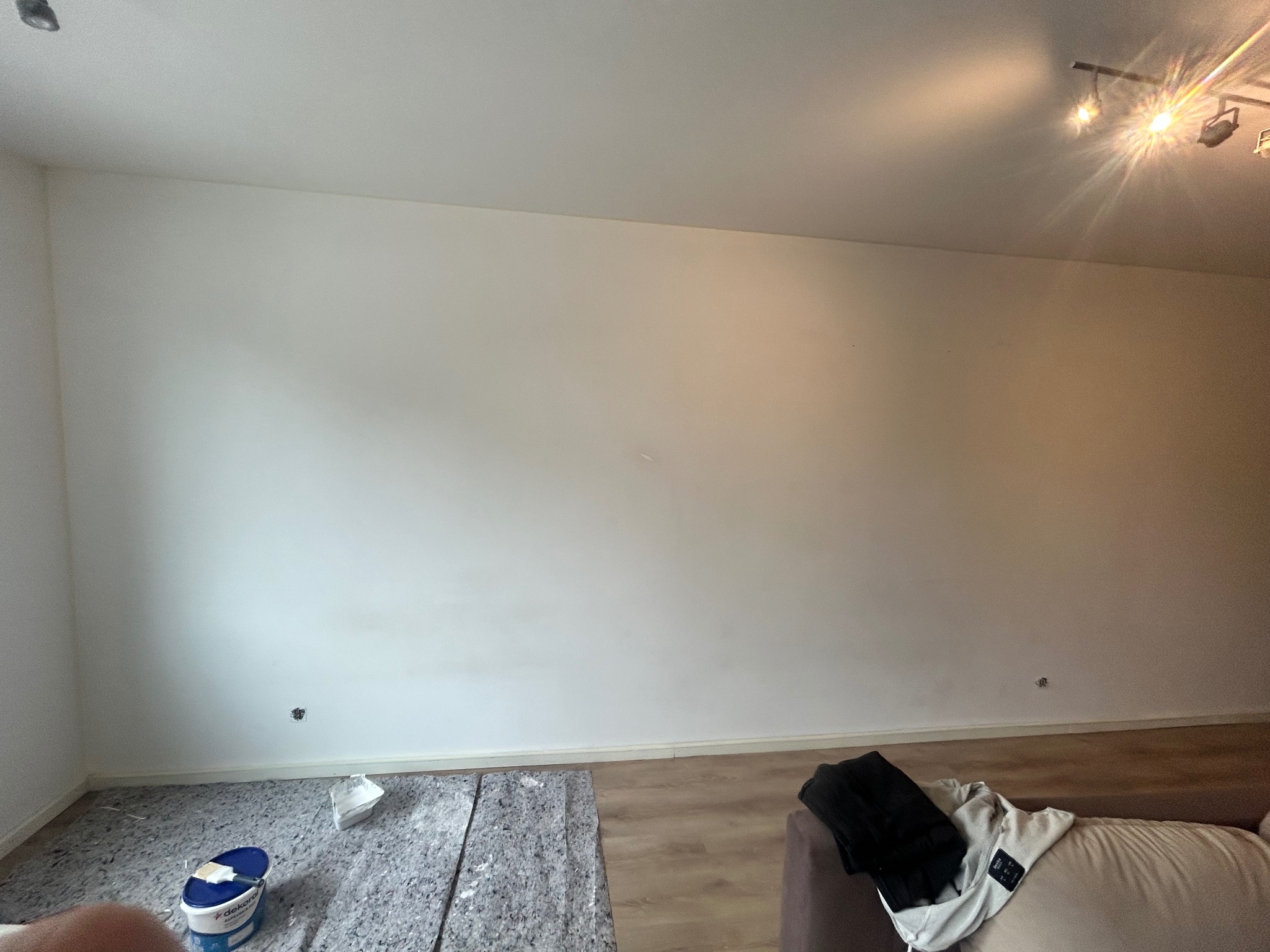 Wnętrze pokoju przed malowaniem: zabezpieczona podłoga, wiadro z farbą w rogu, pusta ściana z widocznymi śladami po gwoździach i lampa sufitowa.