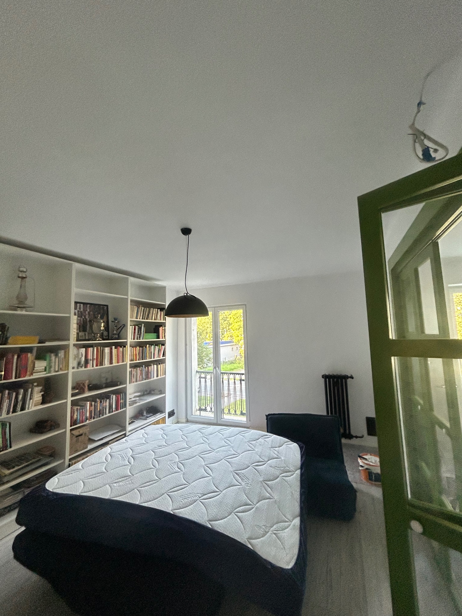 Jasne wnętrze pokoju z regałem na książki, łóżkiem z materacem i widokiem na zieleń za oknem. W tle grzejnik. Wnętrze w trakcie wykańczania.