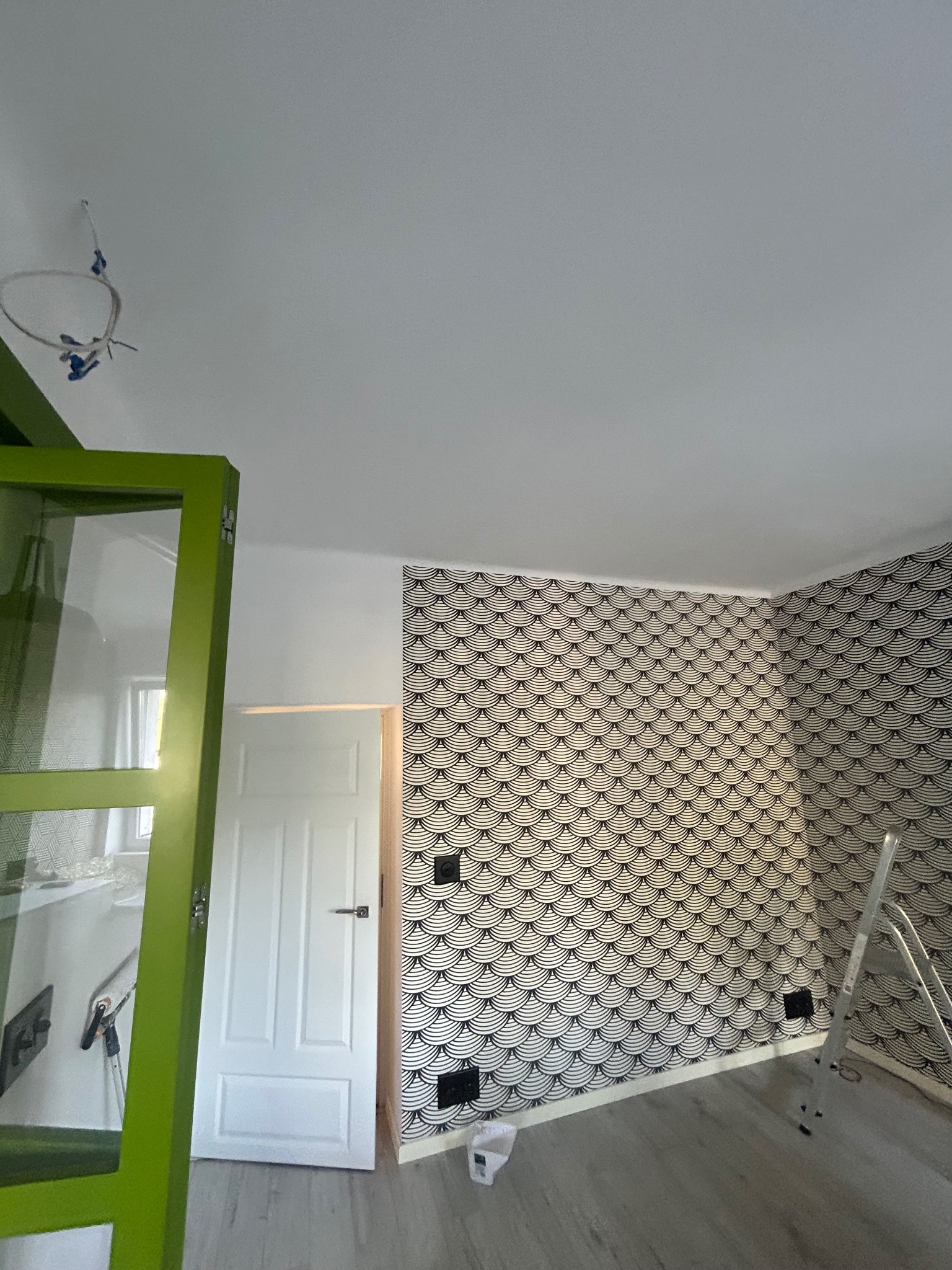 Pokój po tapetowaniu w czarno-białe wzory, drabina oparta o ścianę, widoczny fragment zielonej szafki i białe drzwi. Na suficie wystające kable.