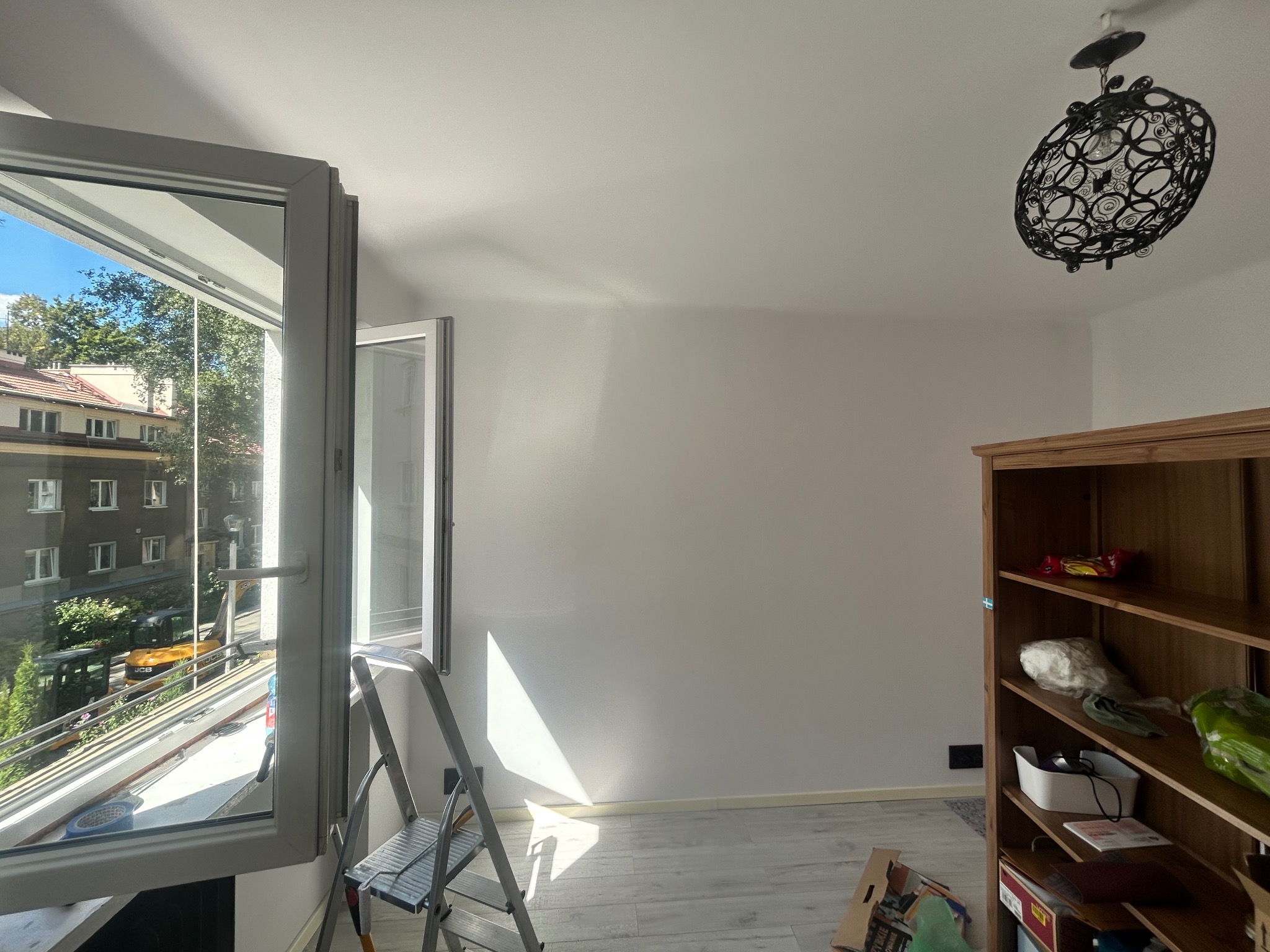 Biała ściana po malowaniu z drabiną i widokiem z okna na zieleń miejską. W rogu drewniany regał i ozdobna lampa sufitowa. Jasne wnętrze po remoncie.