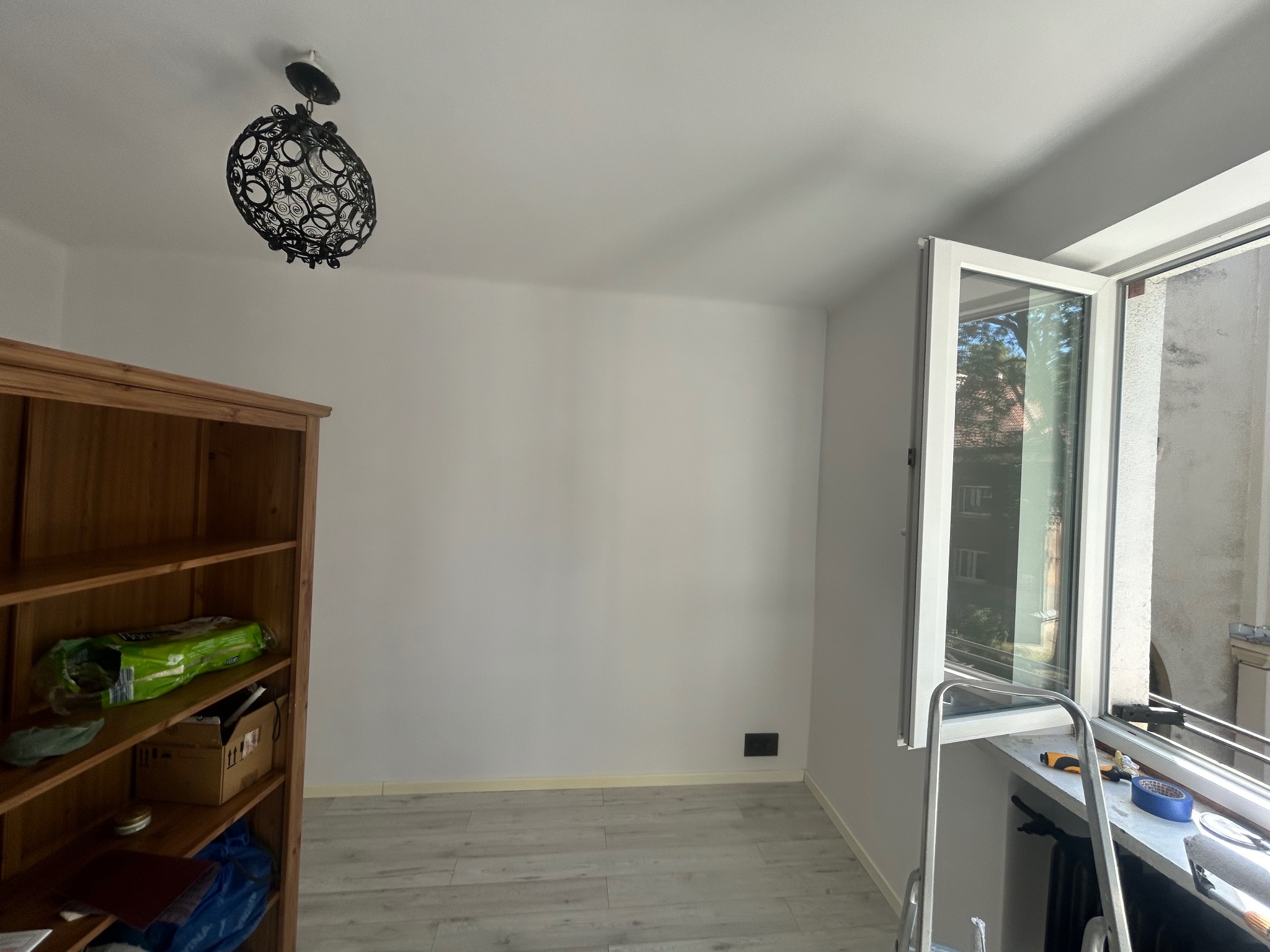 Jasne wnętrze po odświeżeniu: białe ściany, drewniany regał z półkami, otwarte okno z narzędziami na parapecie i drabina. Nowoczesna lampa sufitowa.