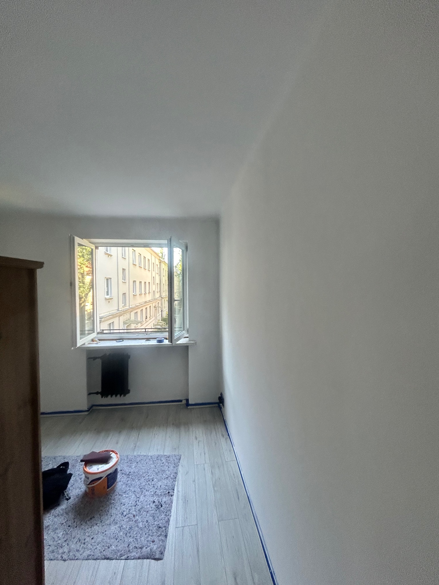 Wnętrze pokoju po malowaniu na biało, z widocznym oknem i grzejnikiem. Na podłodze wiadro z farbą i folia ochronna. Świeżo odmalowane ściany z niebieską taśmą malarską.