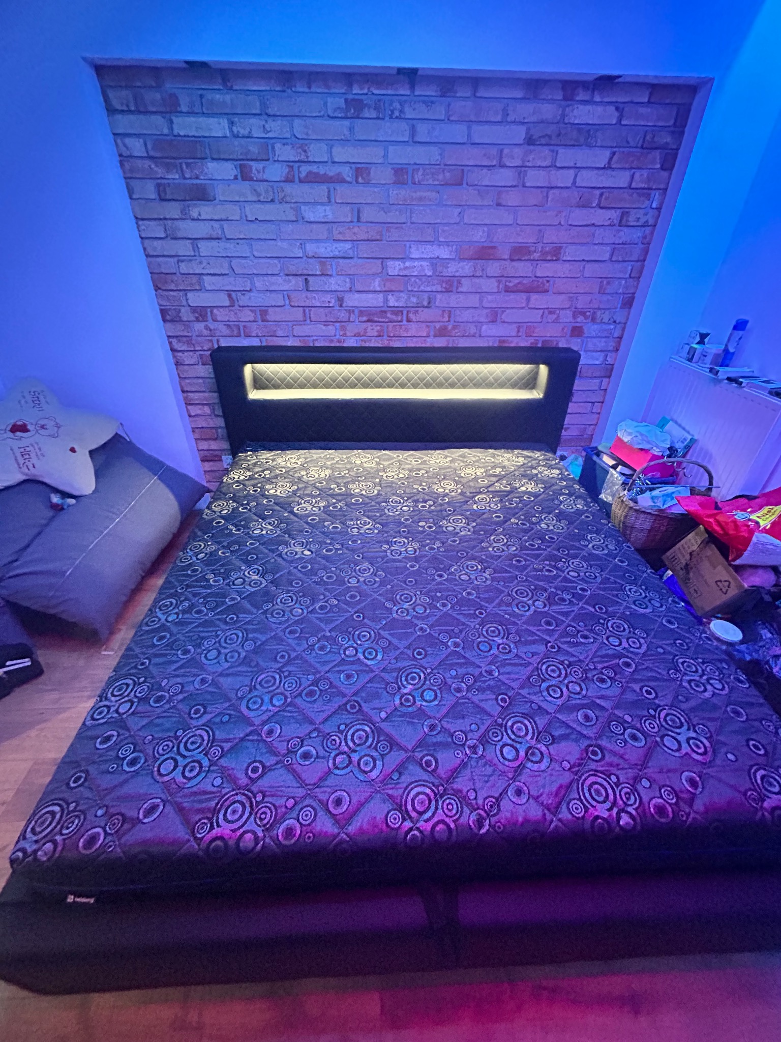 Łóżko z tapicerowanym wezgłowiem i wzorzystą narzutą na tle ściany z cegły, oświetlone niebieskim światłem w pokoju w Krakowie. Ciekawy design.