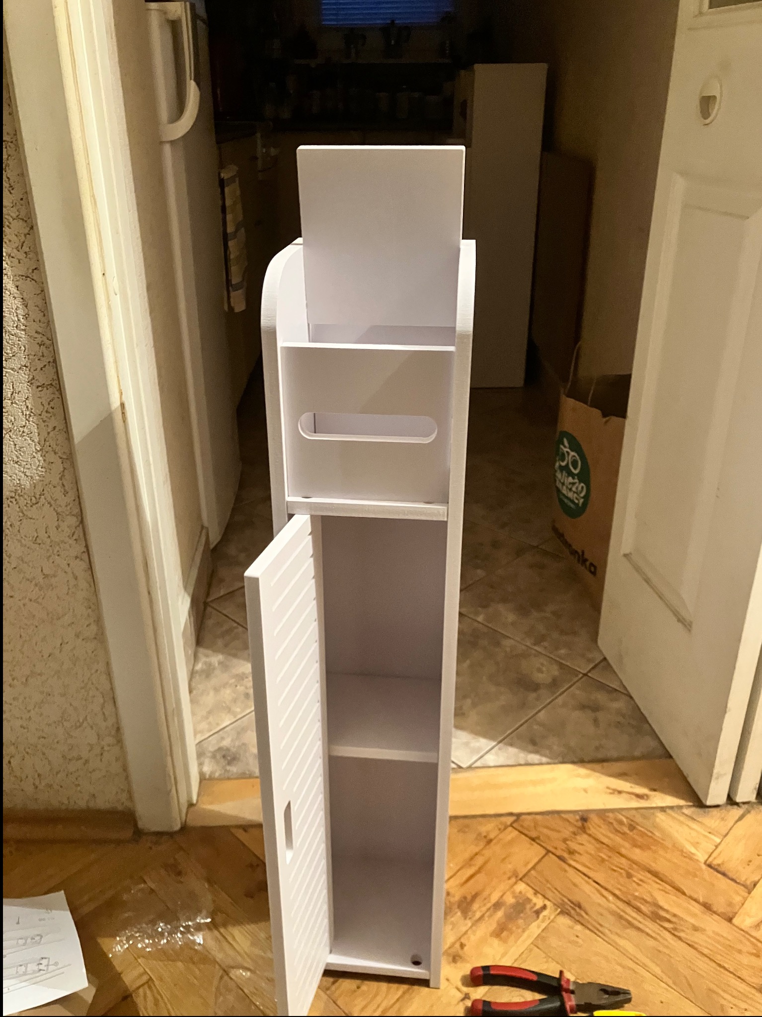 Wąska, biała szafka łazienkowa z otwartymi półkami i uchylnymi drzwiczkami, stoi na parkiecie w pomieszczeniu. Obok leżą kombinerki.