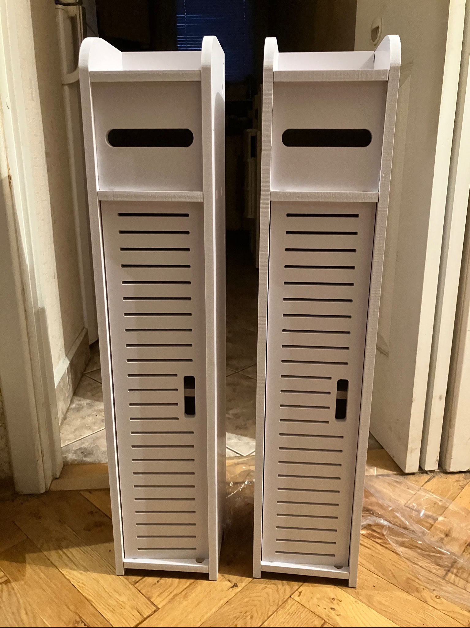 Dwa białe, wąskie regały z lamelkowymi drzwiczkami, ustawione obok siebie na parkiecie. Widoczny fragment futryny drzwi.