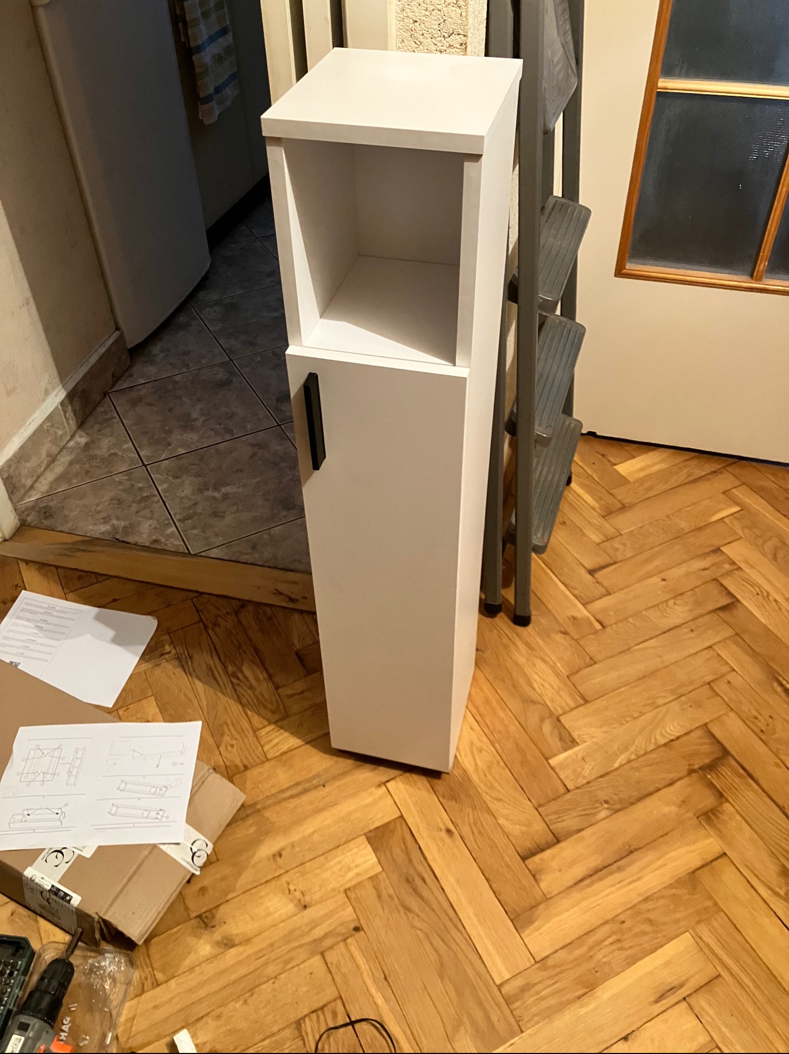 Biała szafka łazienkowa z otwartą półką i drzwiczkami, częściowo złożona, na parkiecie. Obok drabina i kartonowe opakowanie z instrukcją montażu.