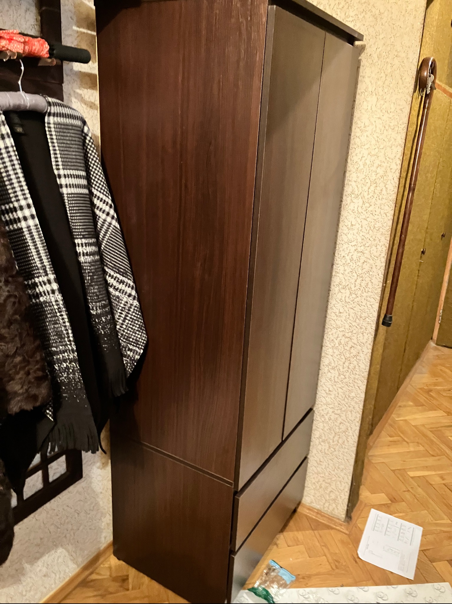Ciemnobrązowa szafa z trzema szufladami, obok wieszak z ubraniami i laska. Na podłodze panele i kartka papieru. W tle ściana z wzorzystą tapetą.