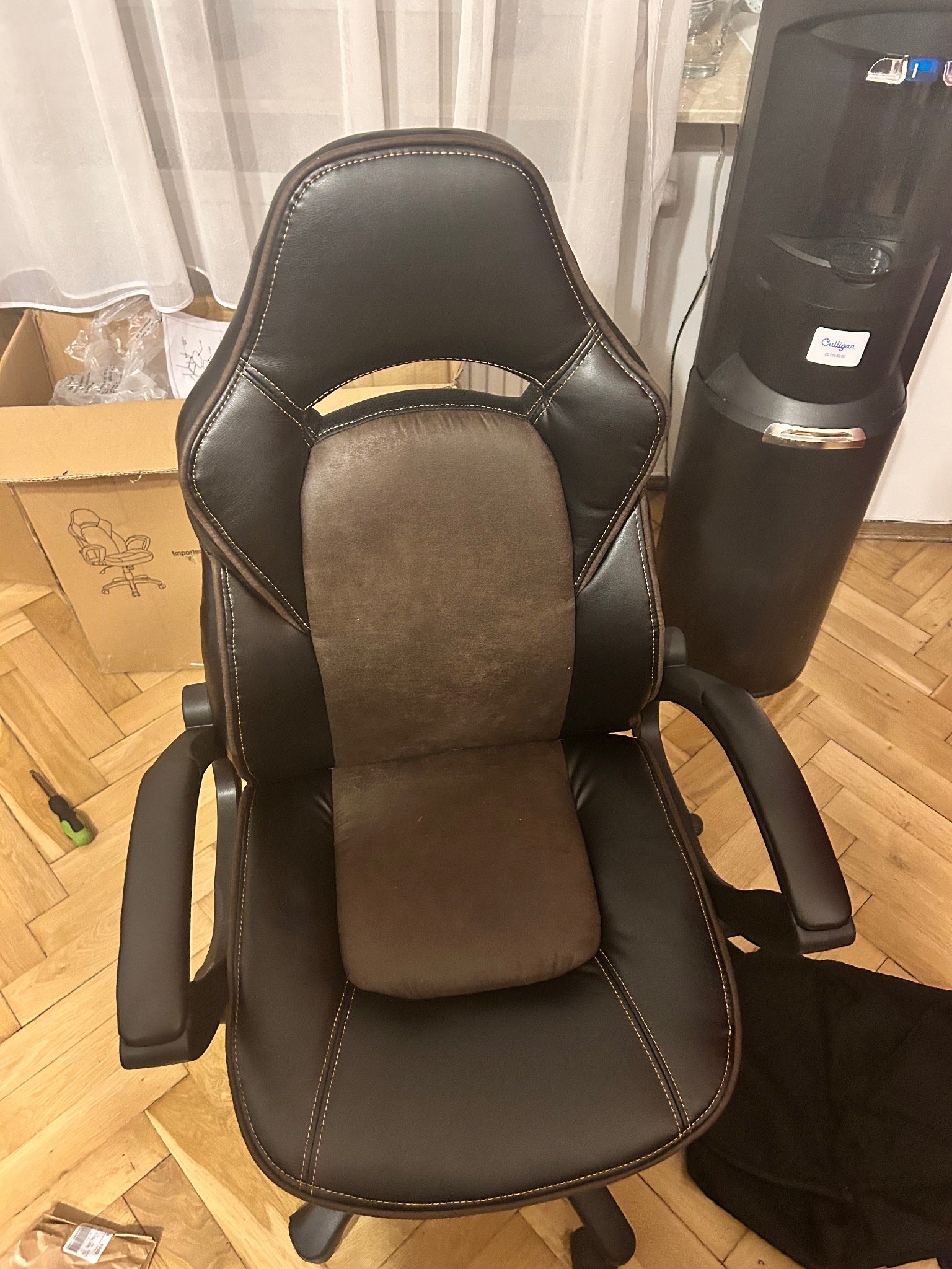 Ciemnobrązowe skórzane krzesło biurowe z brązowym oparciem, widoczne od góry, obok karton i dystrybutor wody. Na podłodze leży śrubokręt i czarny materiał.