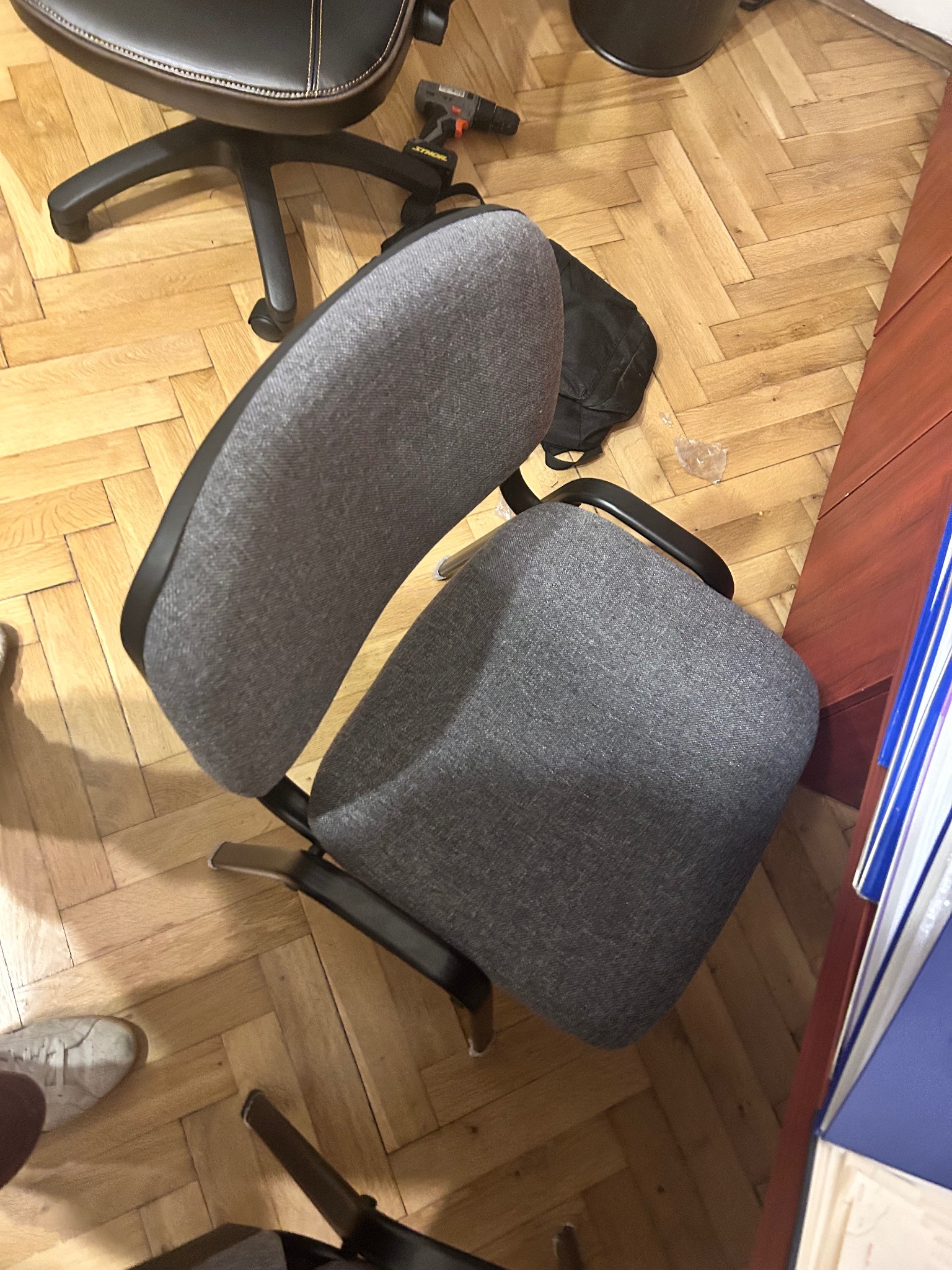 Szary fotel biurowy na kółkach, widok z góry, na parkiecie. Obok wiertarka, torba i fragment biurka z dokumentami. U dołu kadru fragment buta.