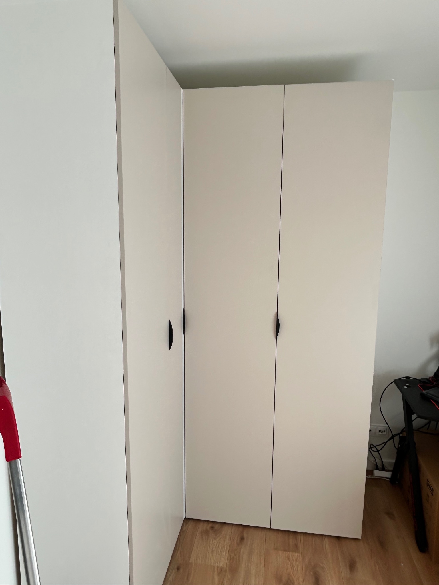 Jasny, narożny segment szafy z trzema drzwiami, z minimalistycznymi uchwytami, ustawiony w rogu pokoju na drewnianej podłodze. Widoczny fragment biurka.