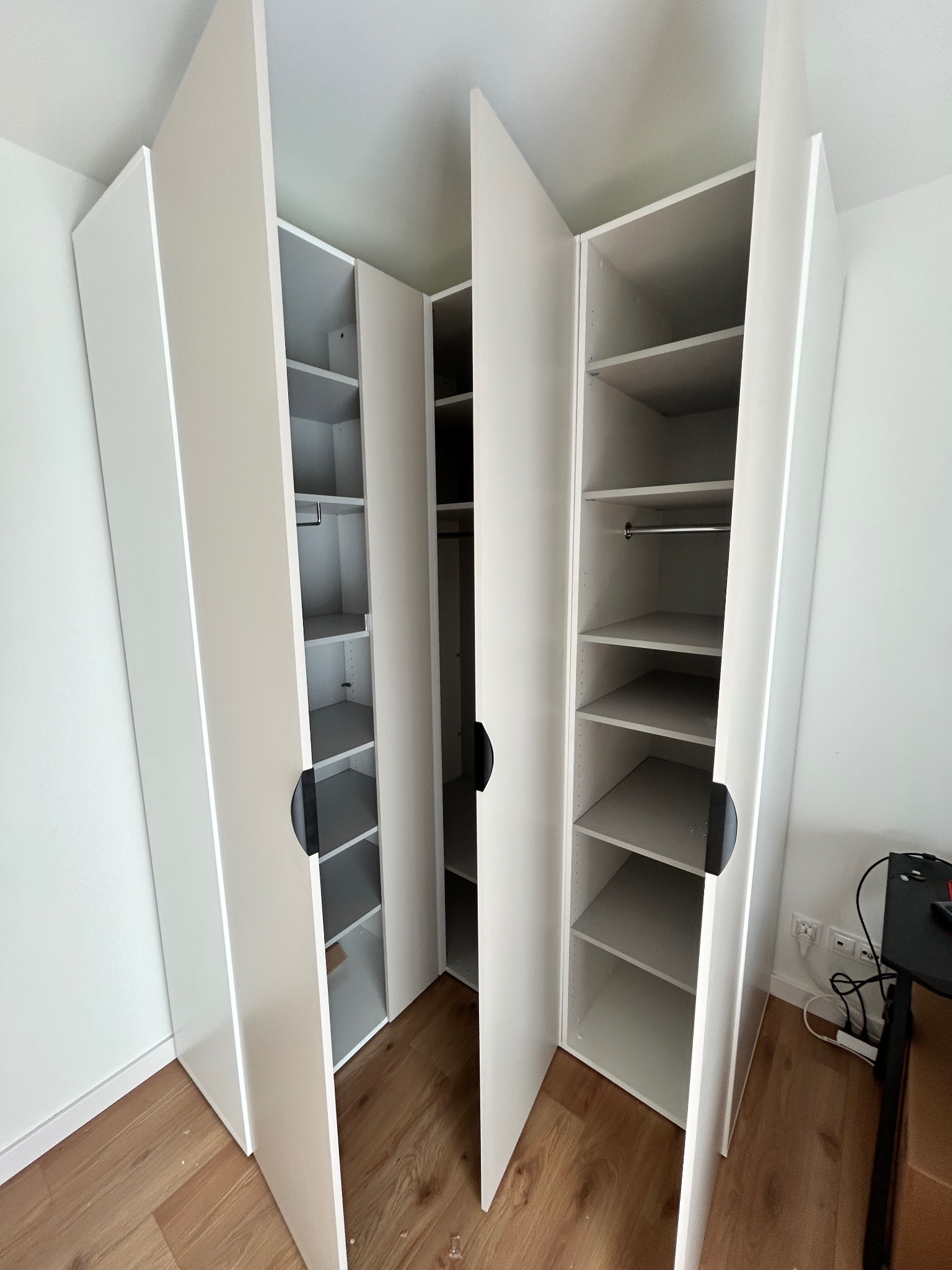 Biała, narożna szafa wnękowa z otwartymi drzwiami, ukazująca półki i drążek na ubrania. Widoczne drewniane panele podłogowe i fragment biurka z elektroniką.
