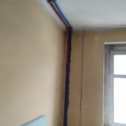 Wewnętrzne instalacje sanitarne: czerwona i niebieska rura poprowadzone wzdłuż ściany pod sufitem, z widocznymi mocowaniami, w pomieszczeniu w trakcie remontu z oknem.