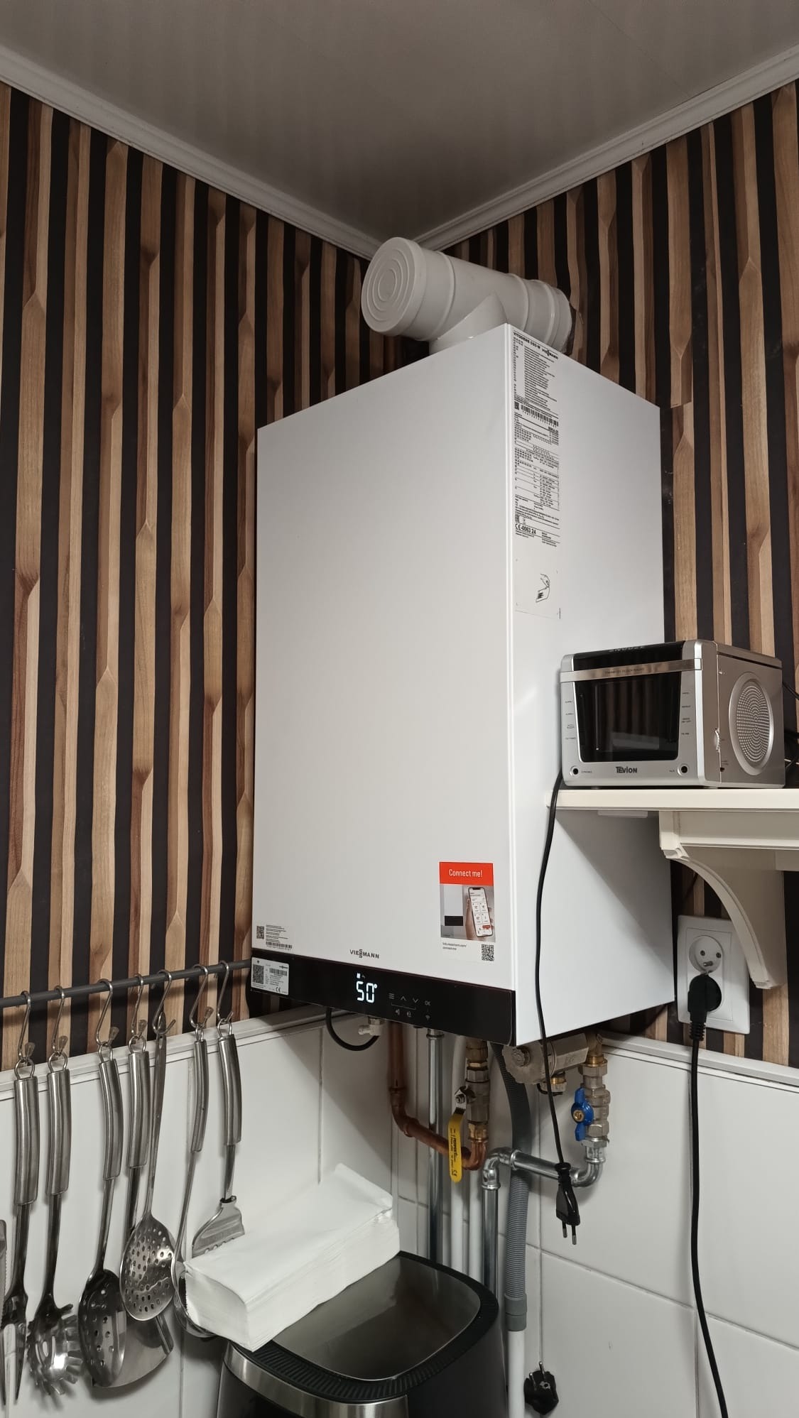 Biały piec gazowy Viessmann z podłączonym kominem, zamontowany na ścianie z dekoracyjną okładziną, widoczne rury i zawory instalacji gazowej, obok półka z małym piekarnikiem elektrycznym.