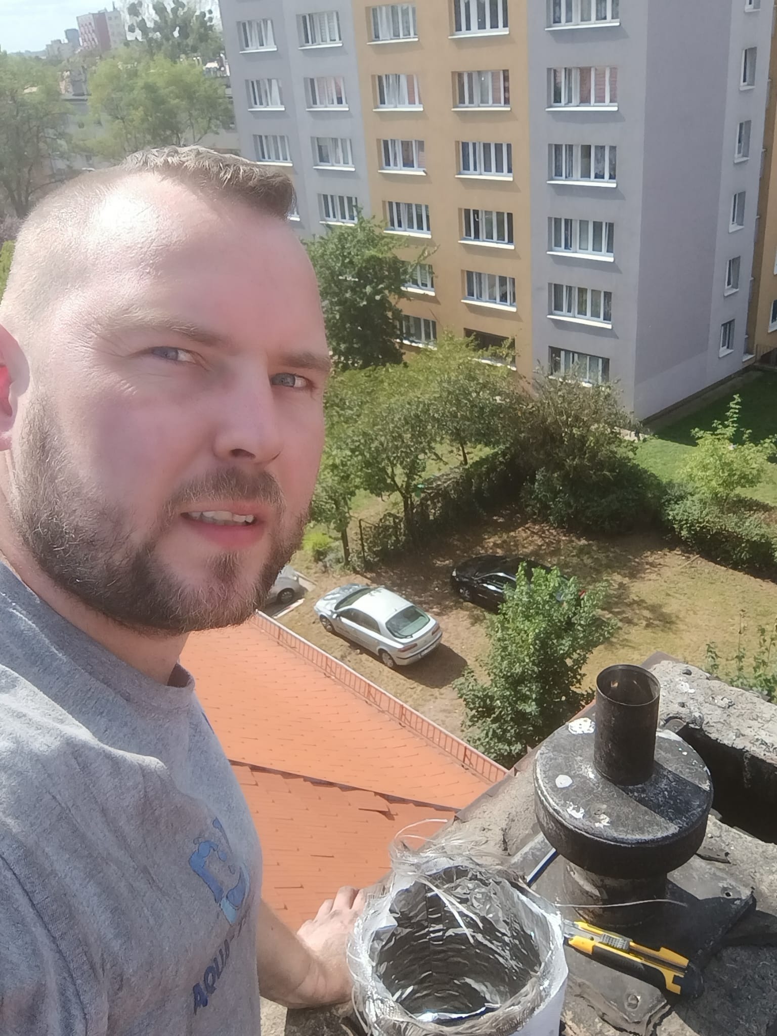 Instalator na dachu budynku mieszkalnego, obok komina z zamontowanym wkładem kominowym, widoczny nóż monterski i fragment instalacji, w tle drzewa i bloki.