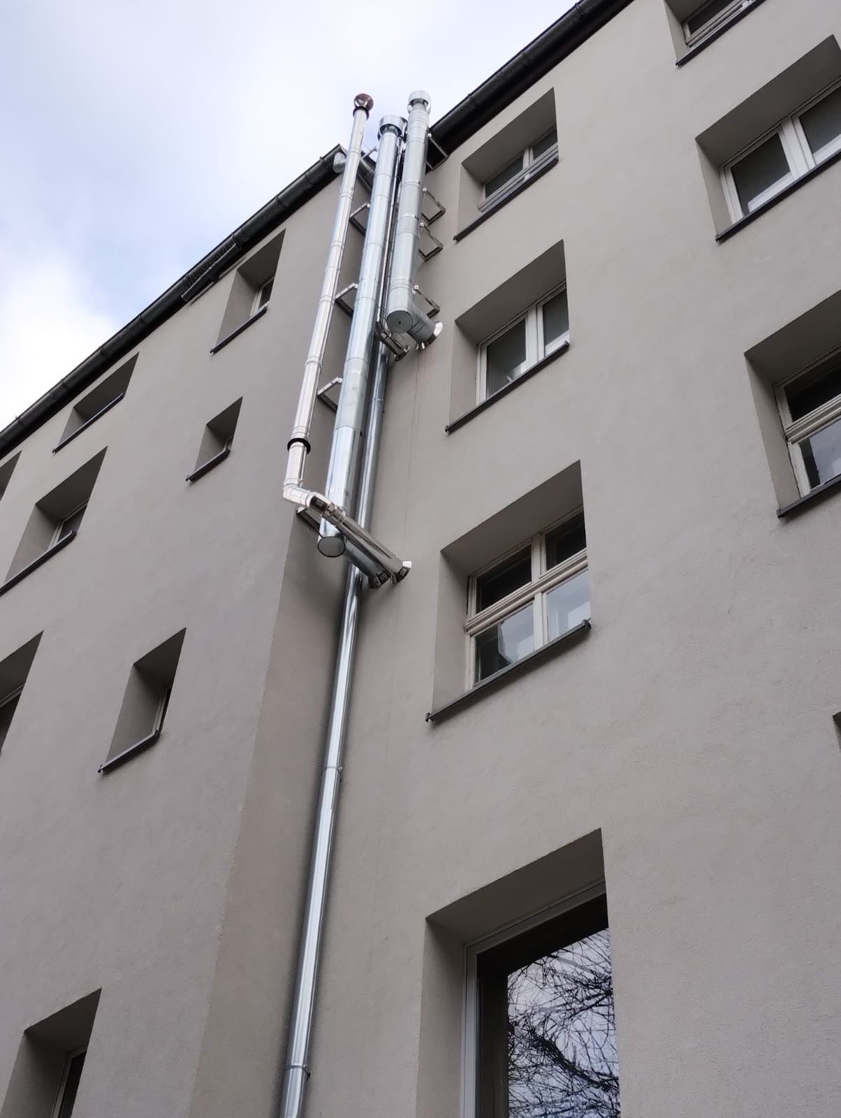 Zewnętrzna instalacja odprowadzania spalin z kotłów gazowych, wykonana z błyszczącej stali nierdzewnej, zamocowana do elewacji budynku mieszkalnego.