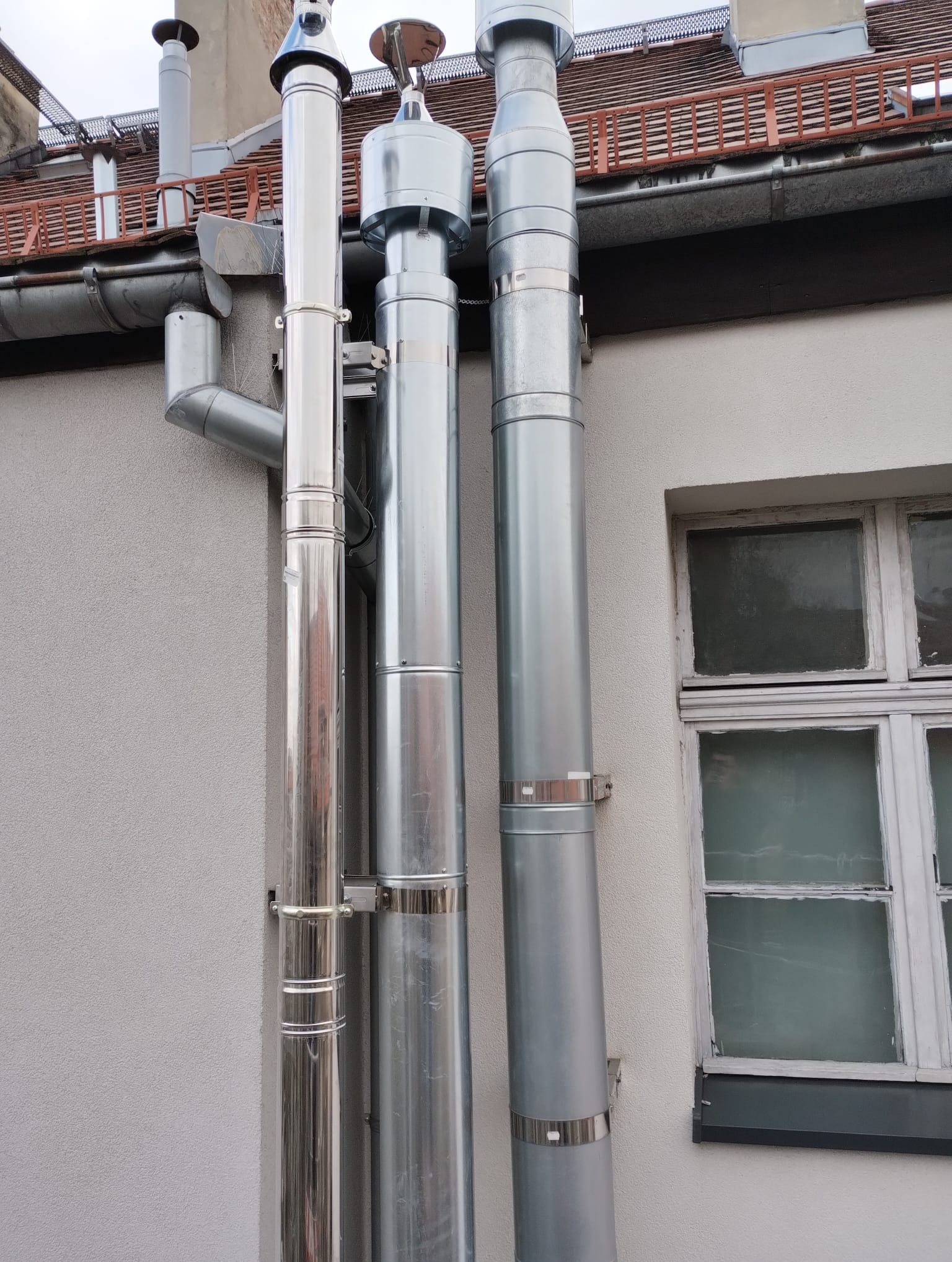 Zewnętrzne kominy spalinowe ze stali nierdzewnej, przymocowane do ściany budynku z widocznym oknem i rynną dachową.
