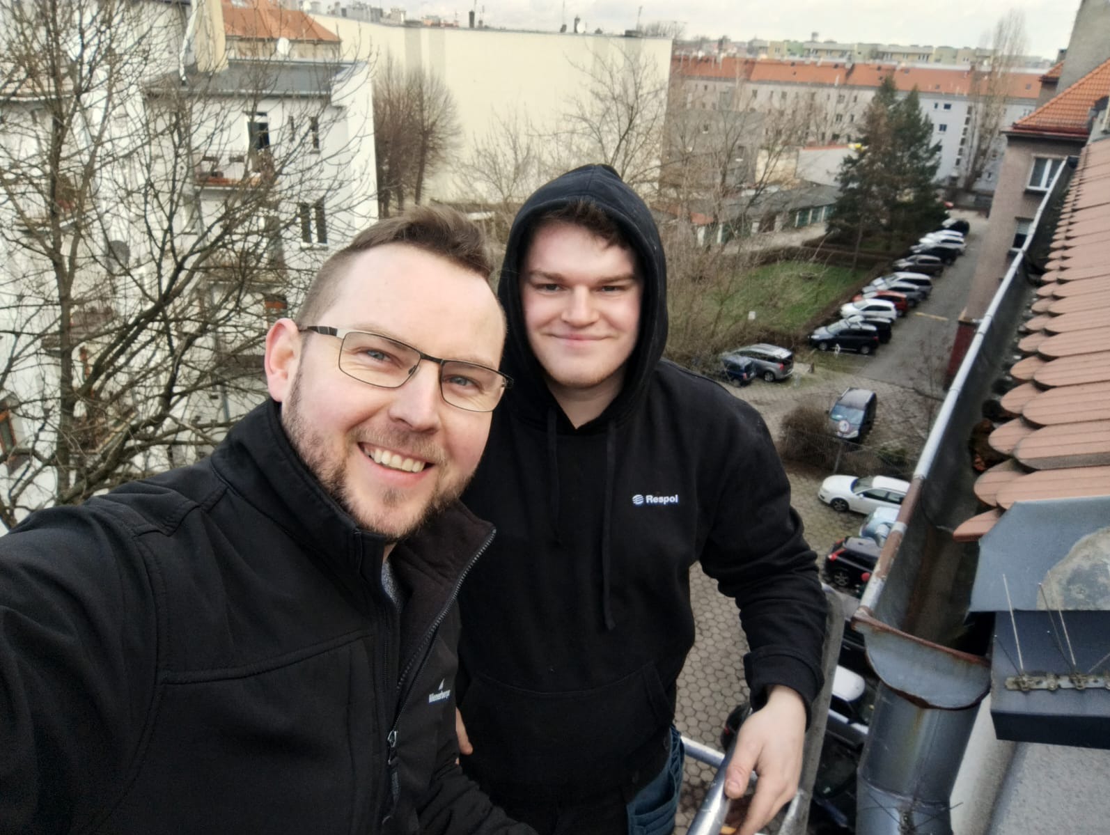 Zdjęcie dwóch uśmiechniętych mężczyzn, prawdopodobnie pracowników firmy Respol, stojących na dachu budynku z widoczną rynną i kolcami przeciw ptakom, z parkingiem i zabudową miejską w tle.