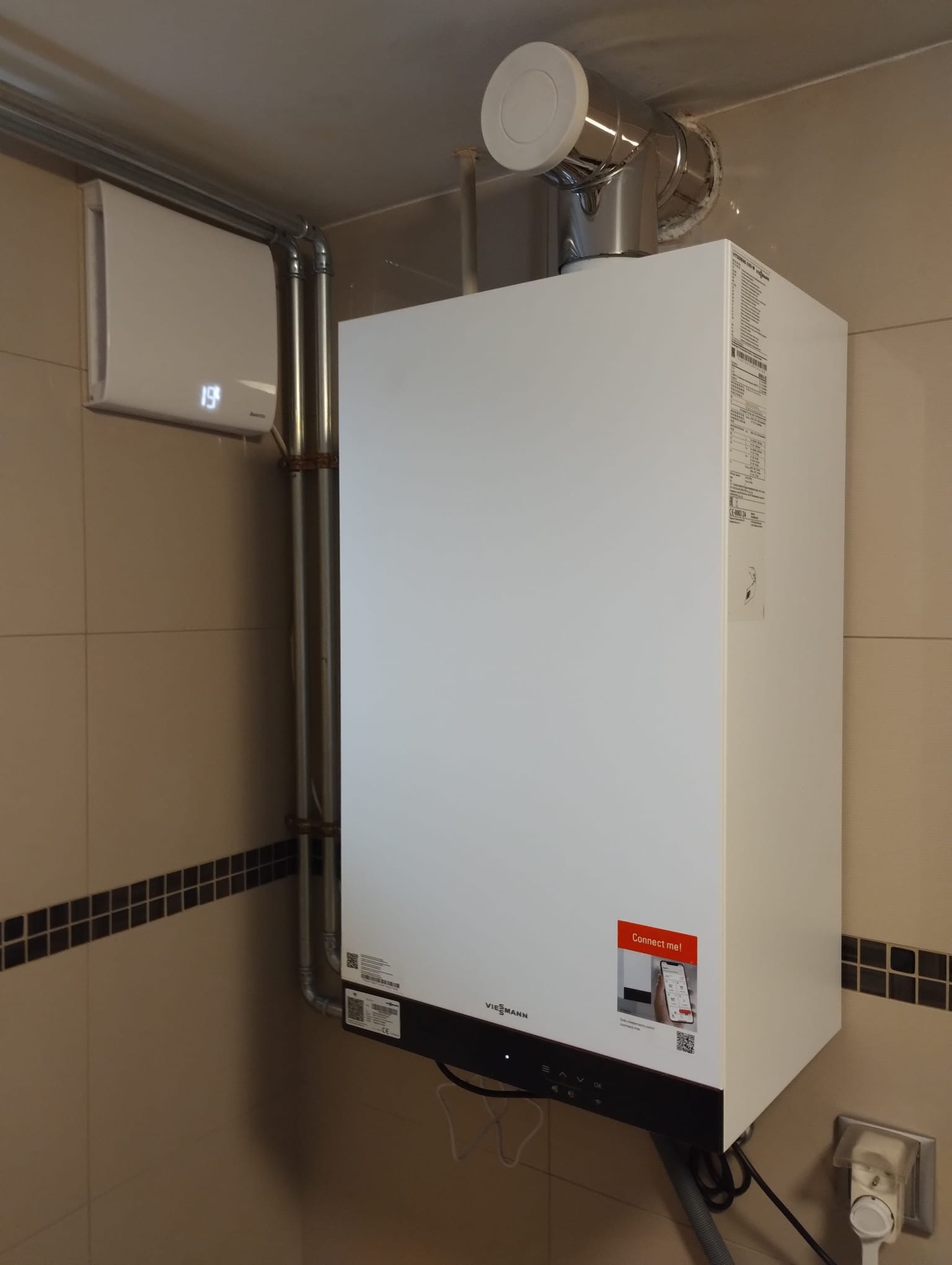 Nowoczesny, biały piec gazowy Viessmann zamontowany na ścianie w łazience z beżowymi płytkami i dekoracyjnym pasem. Widoczne rury odprowadzające spaliny, biały termostat na ścianie wskazujący 19...