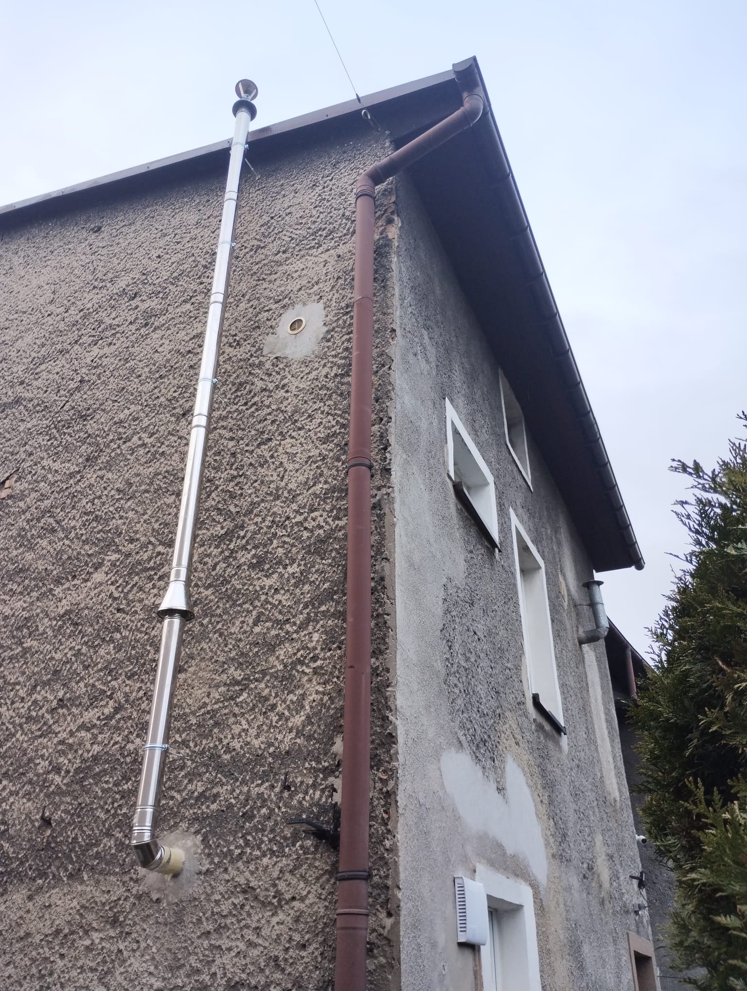 Zewnętrzny, pionowy, stalowy komin spalinowy zamontowany na szarej, teksturowanej ścianie budynku jednorodzinnego, widoczny również brązowy rynna spustowa i fragmenty okien.