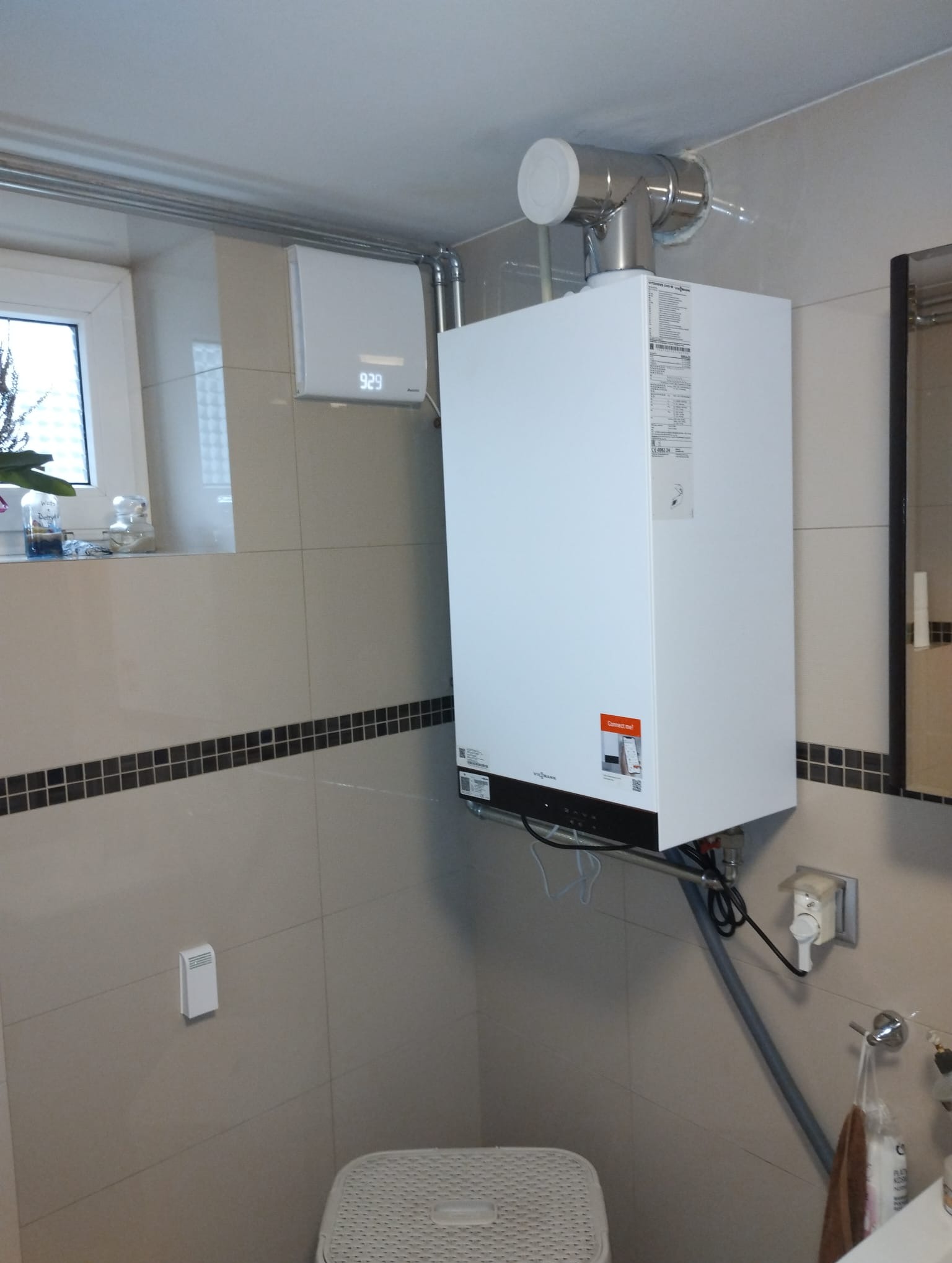 Biały, wiszący piec gazowy Viessmann z podłączonymi rurami w łazience z beżowymi płytkami i dekoracyjnym pasem czarnych kwadratów, widoczny fragment okna z rośliną.