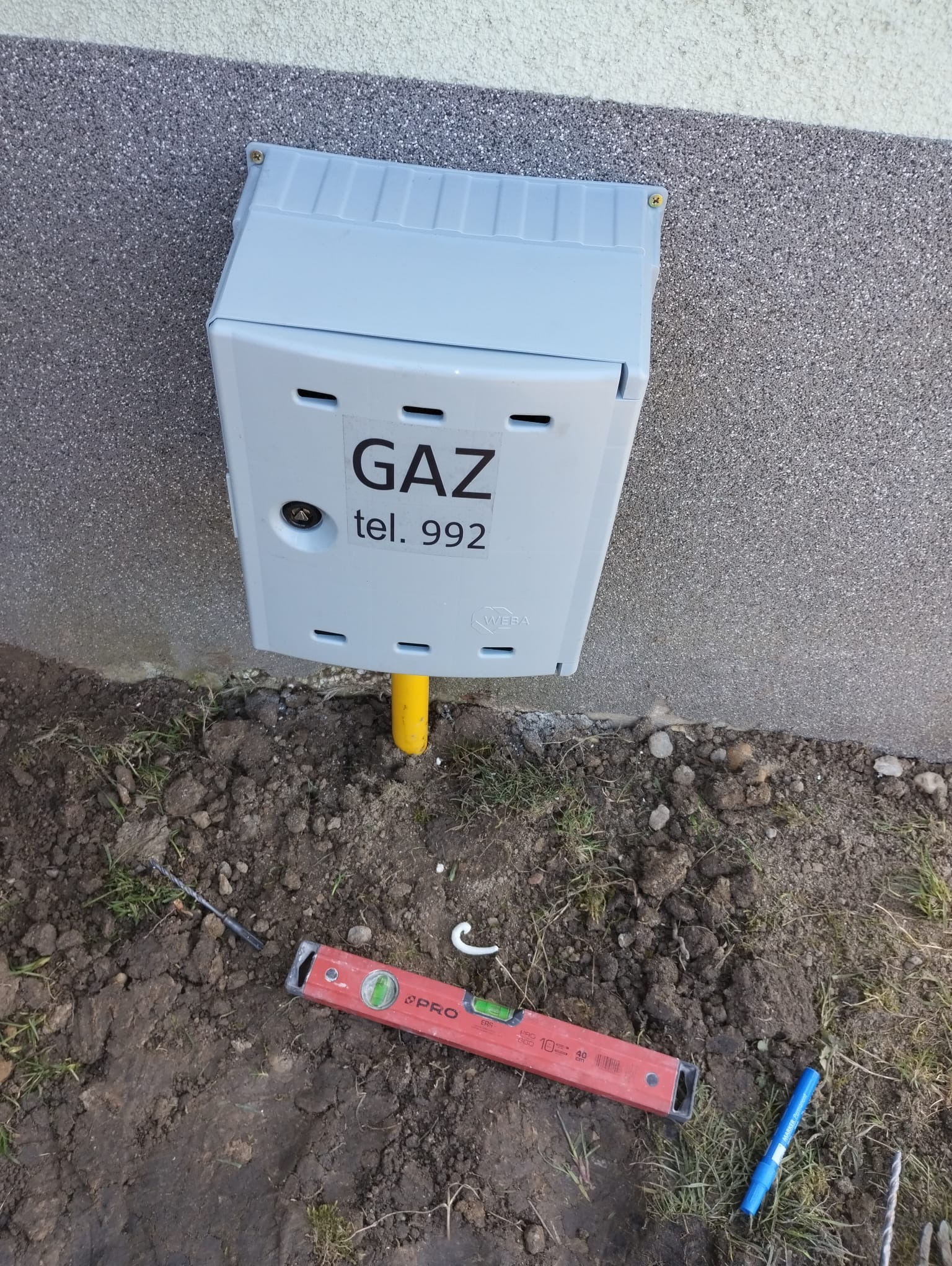 Szara skrzynka gazowa zamontowana na elewacji budynku, z widoczną żółtą rurą, poziomica i wiertło leżące na ziemi.