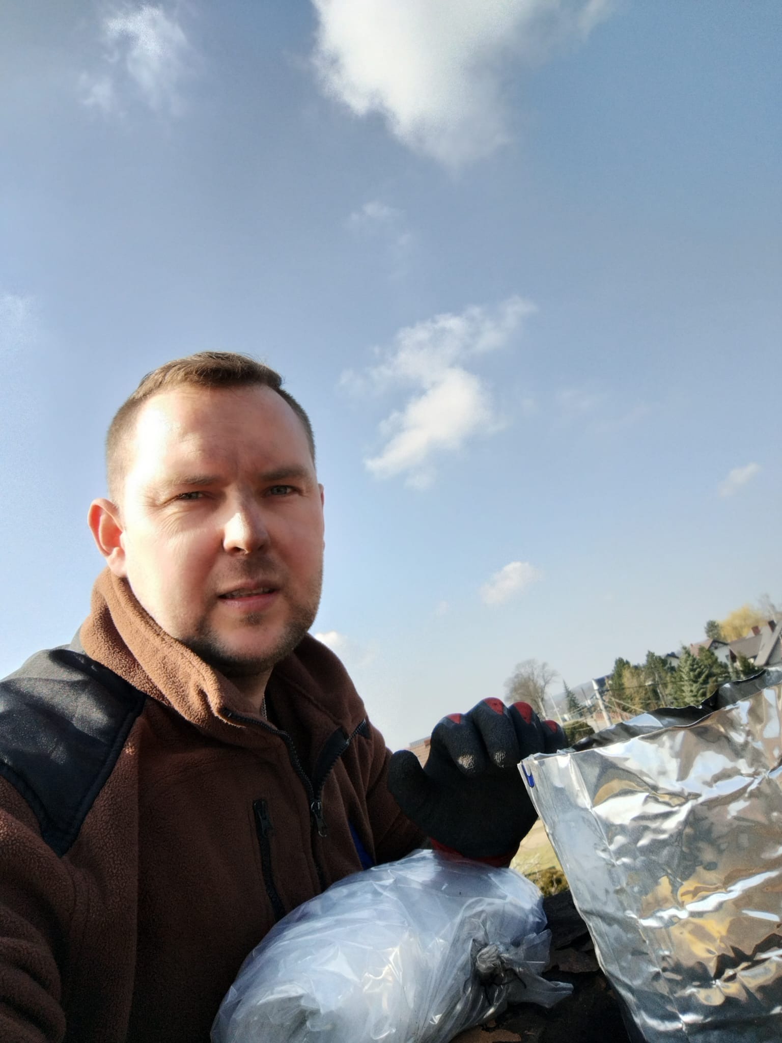 Mężczyzna w roboczych rękawicach trzymający pakunek w folii aluminiowej na tle błękitnego nieba i zabudowań.