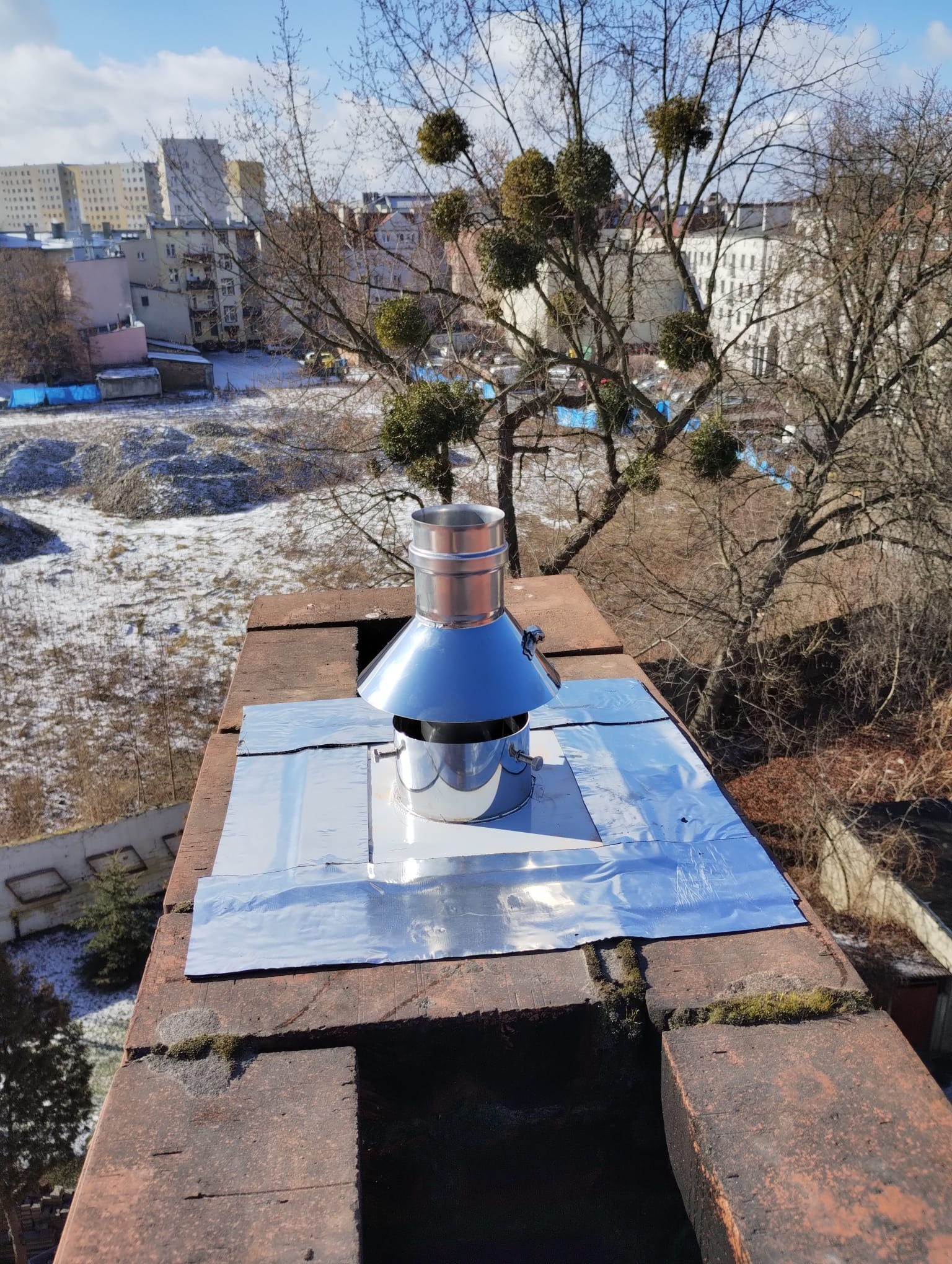 Błyszczący, metalowy komin wentylacyjny na ceglanym kominie, zabezpieczony srebrną folią, w tle widok na zimowy krajobraz miejski i drzewa z jemiołą.