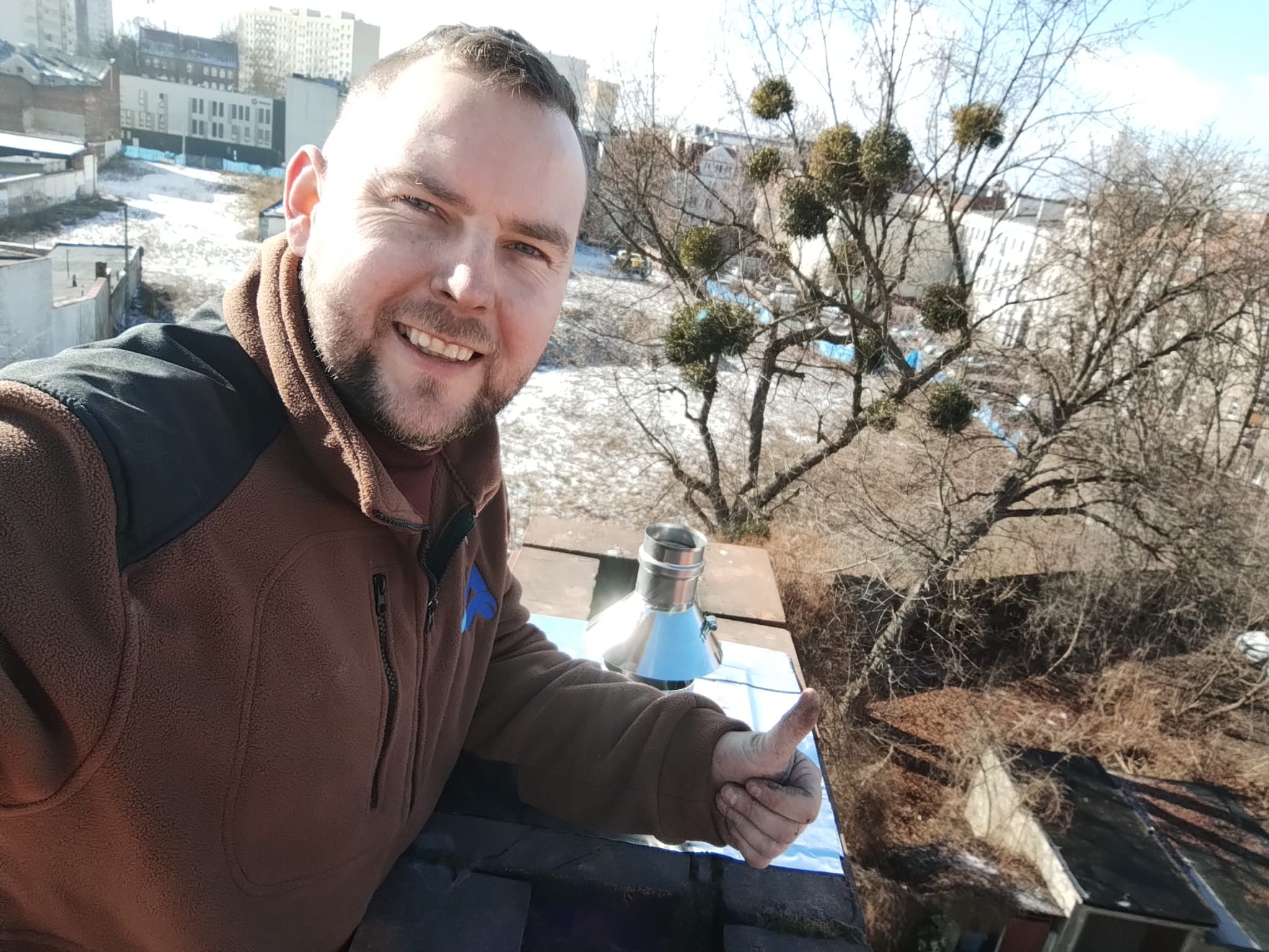 Uśmiechnięty mężczyzna z uniesionym kciukiem na dachu, obok komin z blachy nierdzewnej, w tle budynki i drzewa z jemiołą.