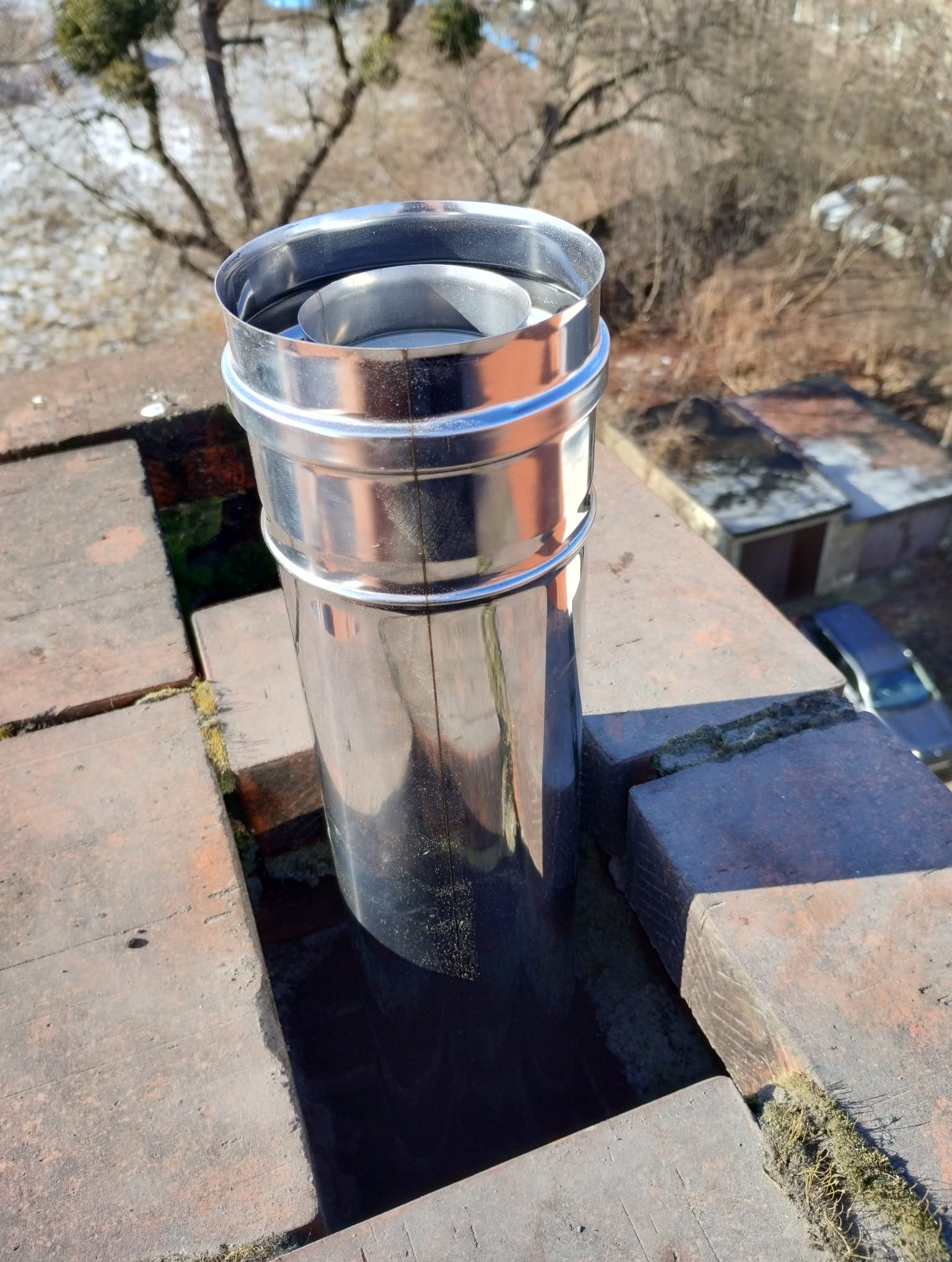 Błyszczący, metalowy komin na ceglanym dachu z mchem, widok z góry w słoneczny dzień.