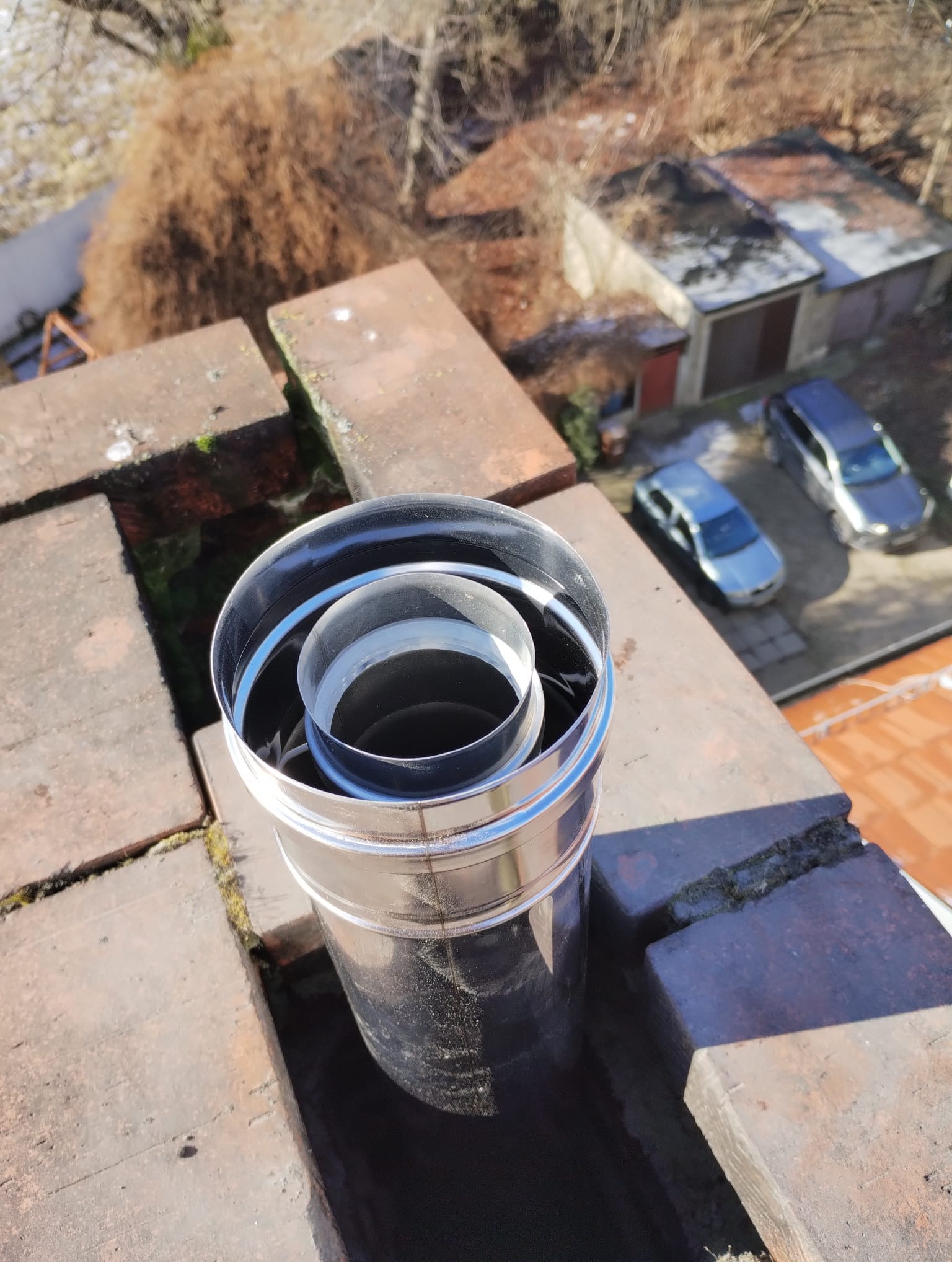 Komin wentylacyjny z rur stalowych na dachu z cegły, widok z góry na otoczenie z budynkami i samochodami.