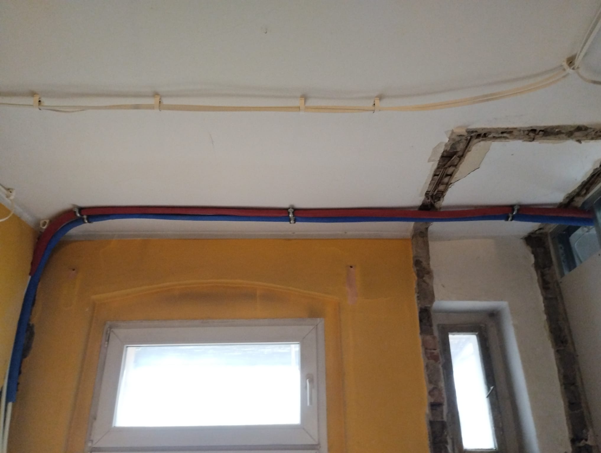 Dwie rury, czerwona i niebieska, przymocowane do ściany i sufitu za pomocą metalowych uchwytów, widoczne na tle żółtej ściany z oknem i fragmentu sufitu z widoczną konstrukcją.