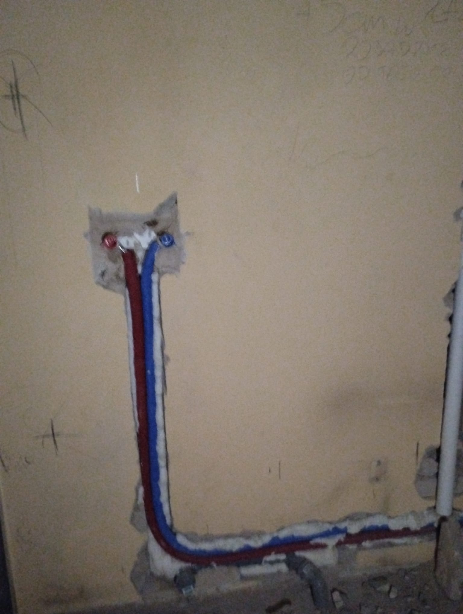 W ścianie widoczne rury instalacji wodnej (czerwona i niebieska) w trakcie montażu, zabezpieczone pianką montażową, z widocznymi oznaczeniami na ścianie.