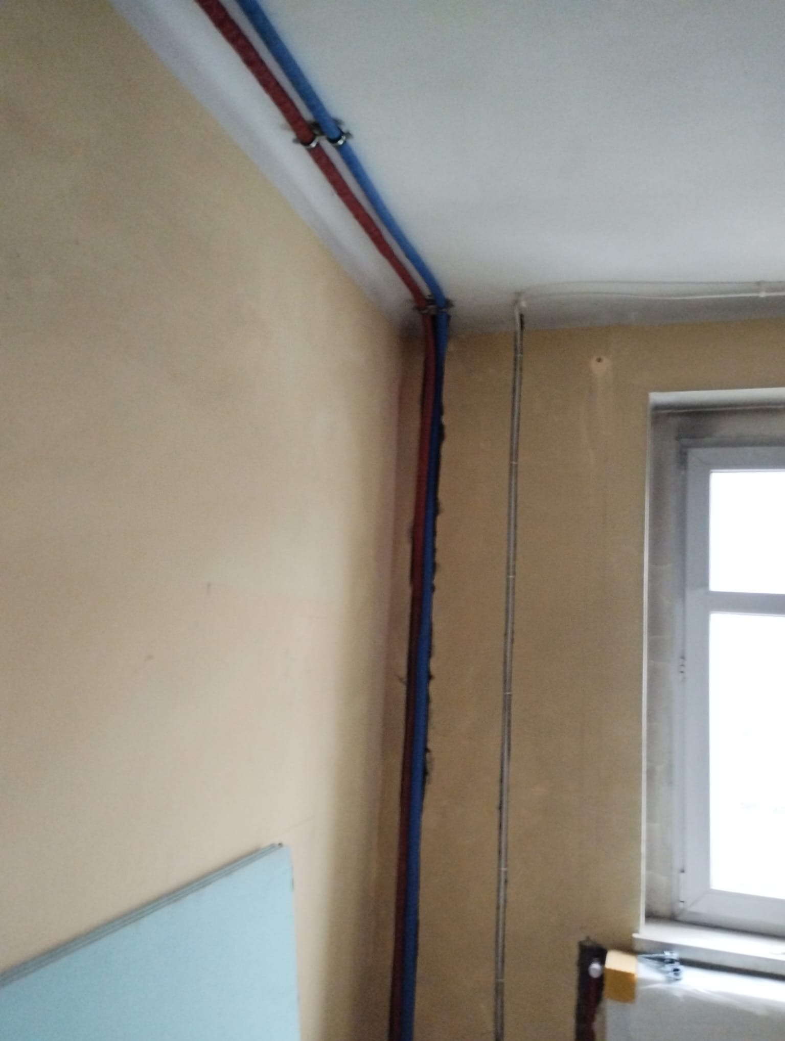 Wewnętrzne instalacje sanitarne: czerwona i niebieska rura poprowadzone wzdłuż ściany pod sufitem, z widocznymi mocowaniami, w pomieszczeniu w trakcie remontu z oknem.
