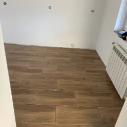 Brązowe płytki imitujące drewno ułożone na podłodze w pomieszczeniu w trakcie remontu, widoczne przygotowane punkty elektryczne na ścianie i grzejnik.